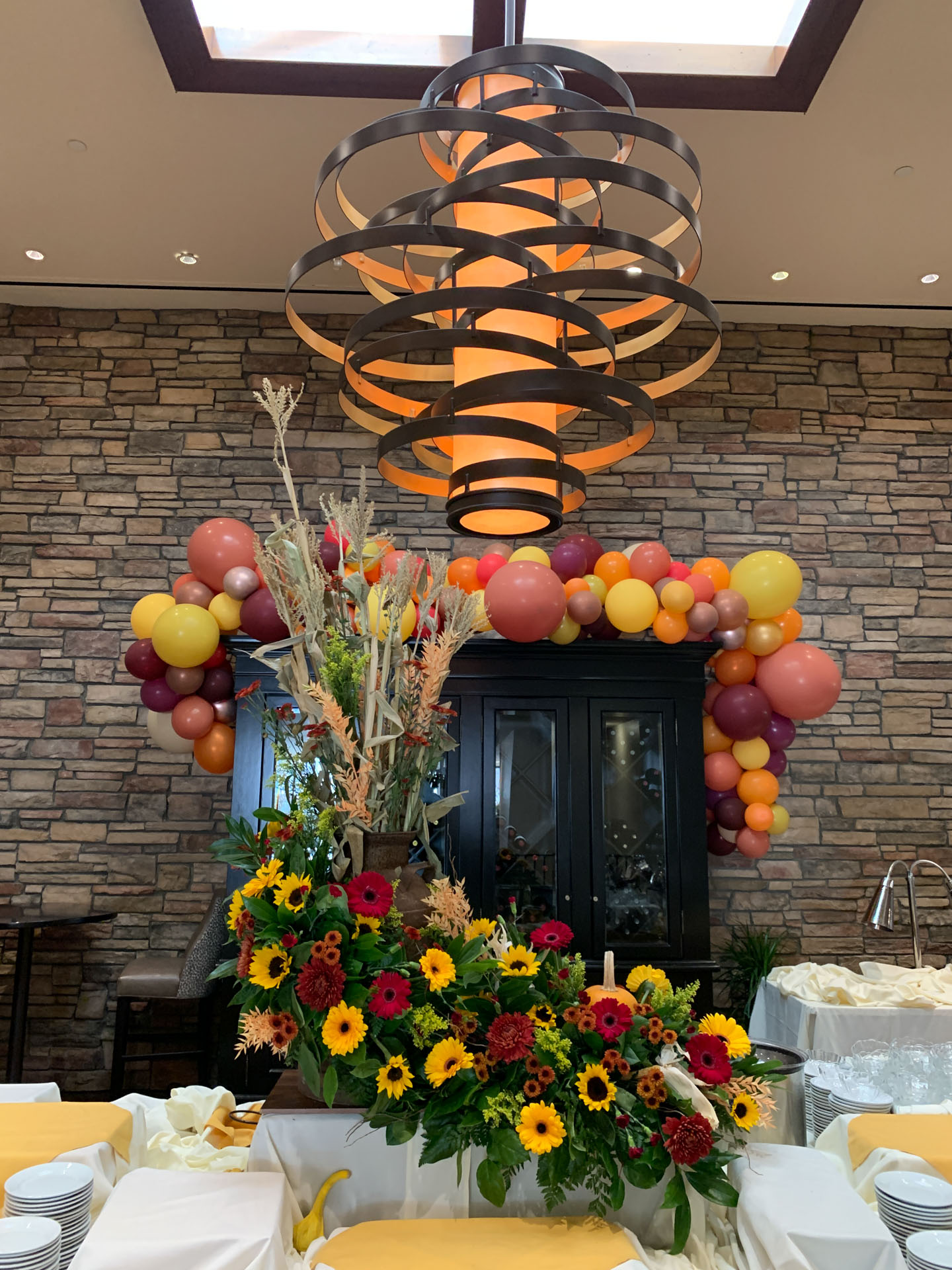 Indoor floral and balloon decorations with red, yellow, orange flowers and balloons on black cabinet under a large abstract oval chandelier.