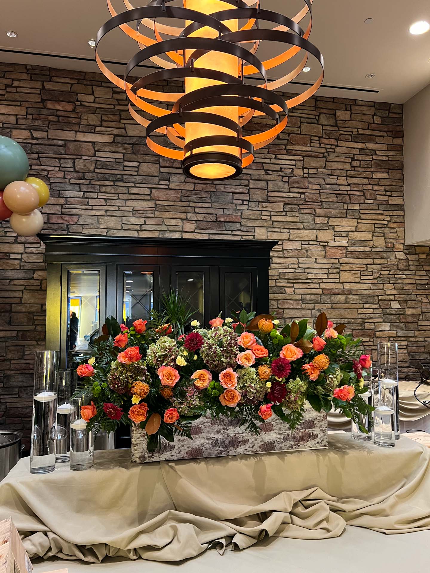 Ornate floral arrangement with orange, red, and green flowers on a draped beige cloth table under an abstract chandelier.