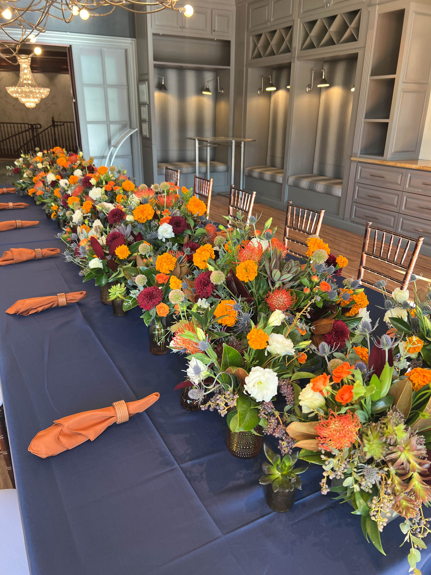 Long table with orange and white floral centerpieces and orange napkins on a dark blue tablecloth.