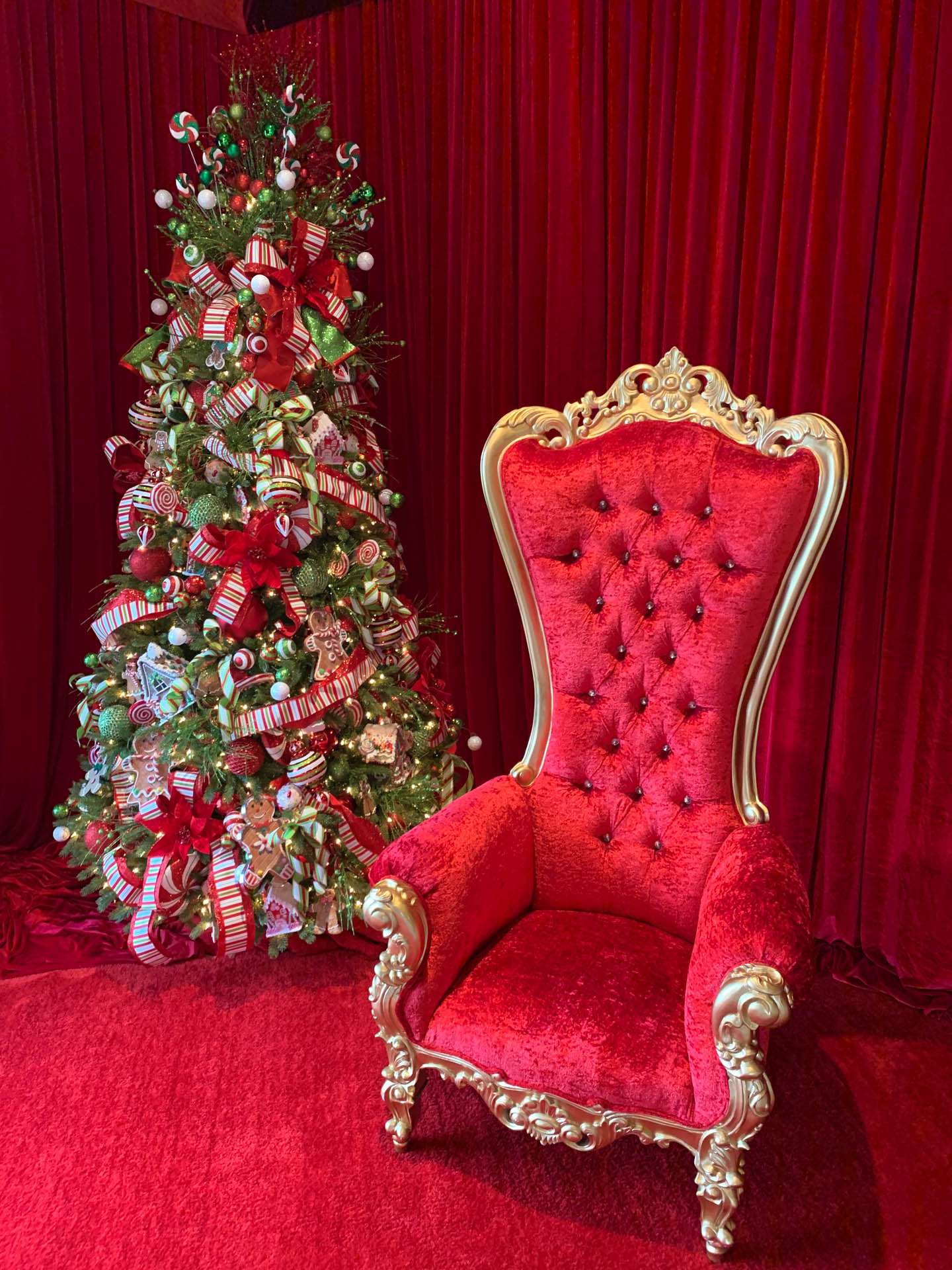 Red velvet Christmas tree decorated with candy and gingerbread beside ornate red and gold armchair.