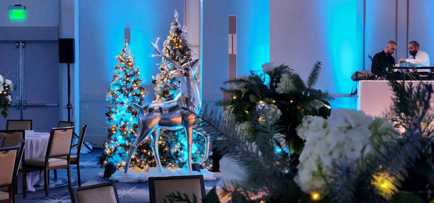 Indoor event space decorated with lit Christmas trees, a shiny reindeer statue, and floral arrangements with a snowy winter theme.