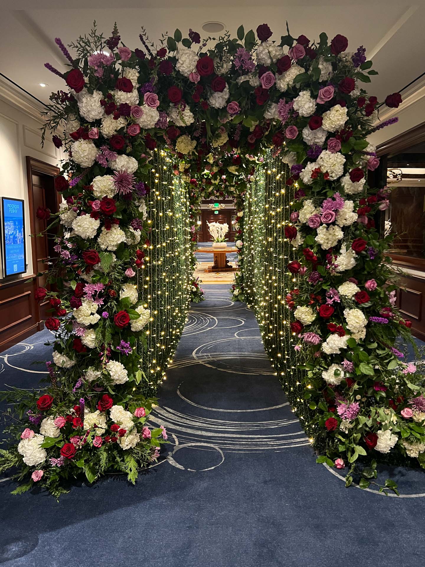 Indoor floral archway decorated with red, white, and purple flowers and string lights on blue carpet.