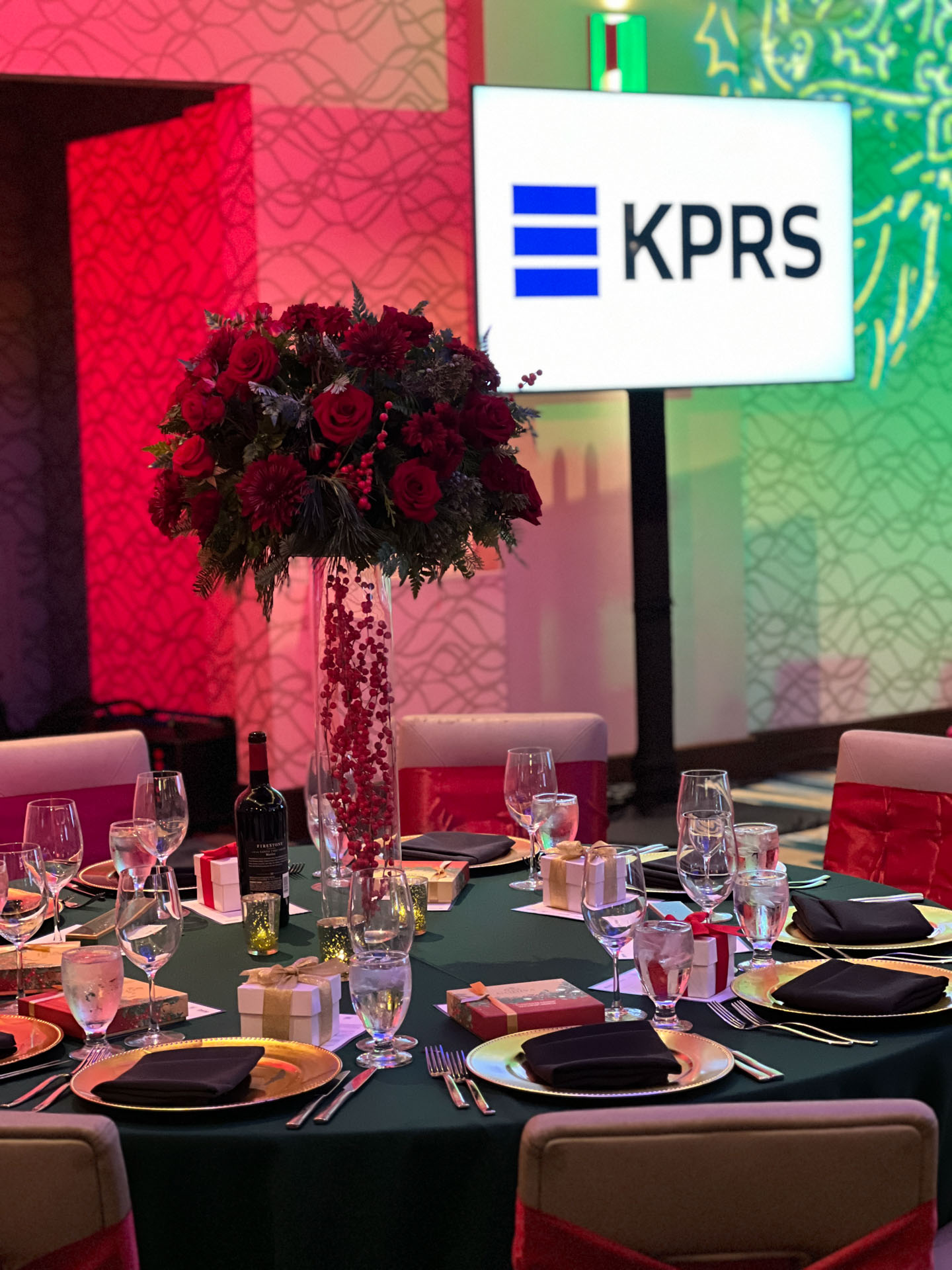 Round table set for a snowy winter Christmas themed event with gold plates, black napkins, glasses, gift boxes, and a tall red floral centerpiece.