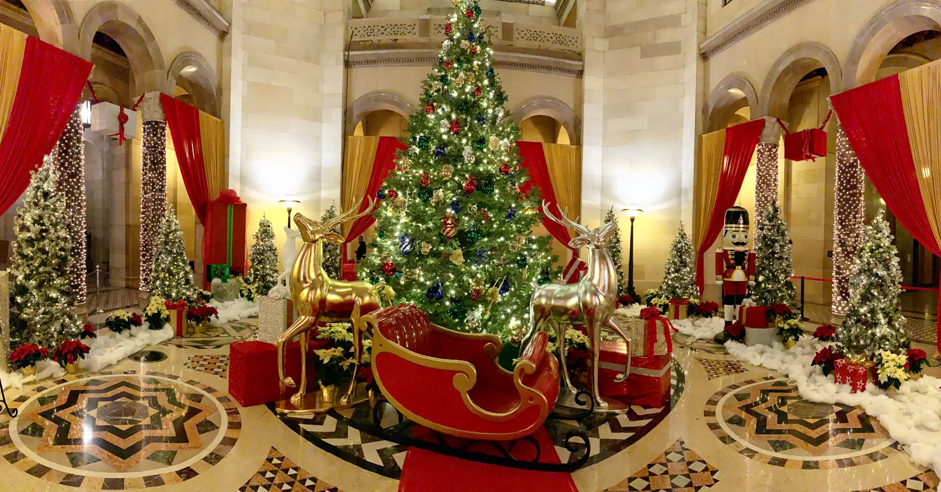 Decorated Christmas tree with lights, gold and silver reindeer, red sleigh, and a nutcracker in a grand hall.