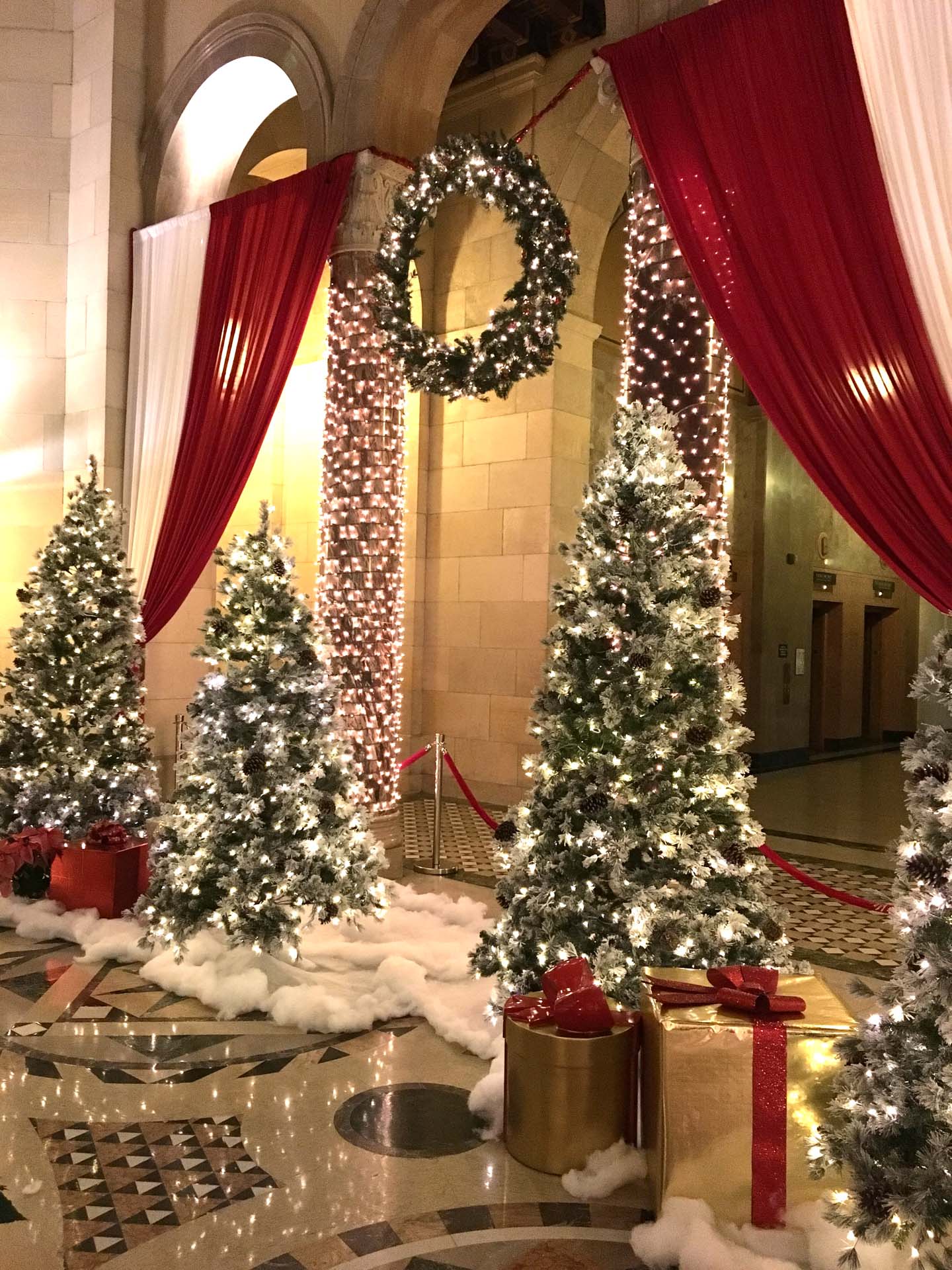 Christmas trees with white lights and pinecones, red and gold wrapped gifts, and a lit wreath in a grand hall.
