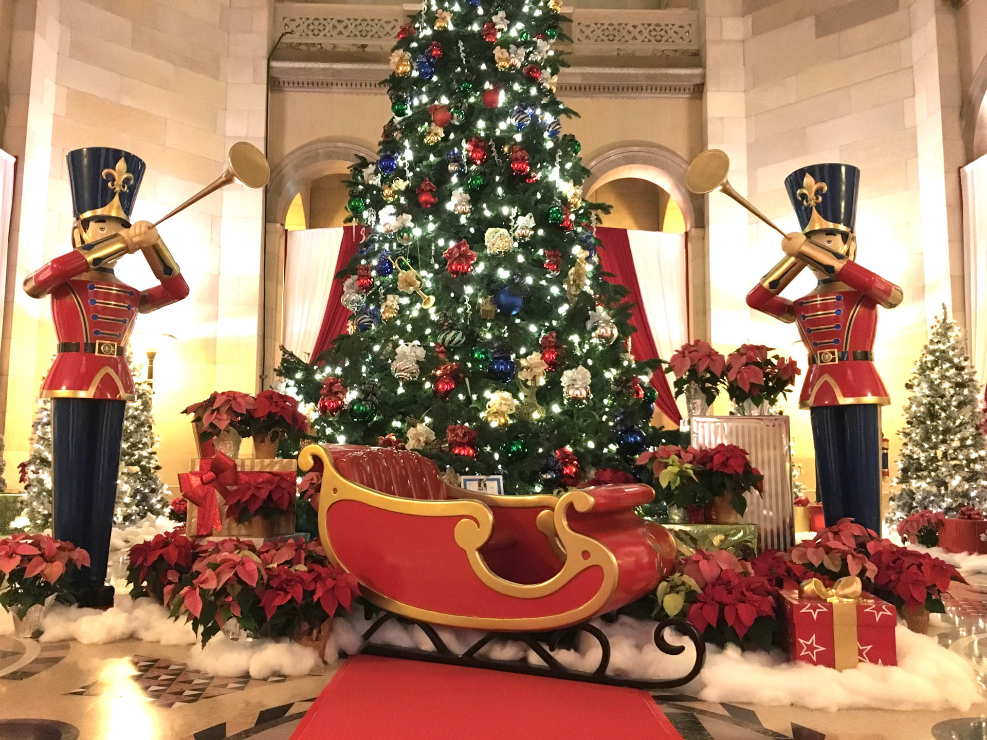 Christmas tree decorated with lights and ornaments, flanked by two toy soldier hornblower statues and a red and gold sleigh.