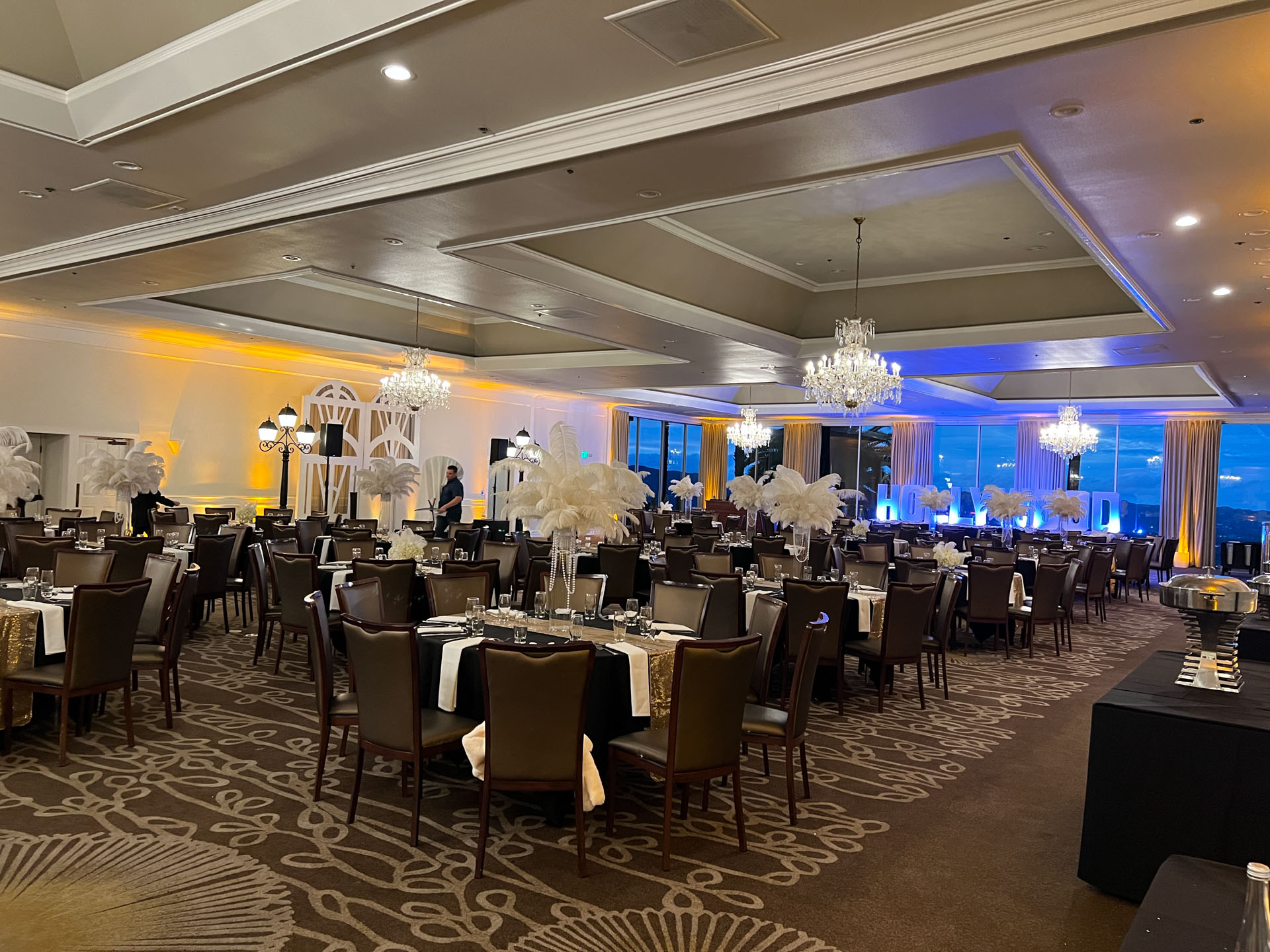 Elegant banquet hall with chandeliers, decorated tables, and 'HOLLYWOOD' letters lit with up lights against a large window.