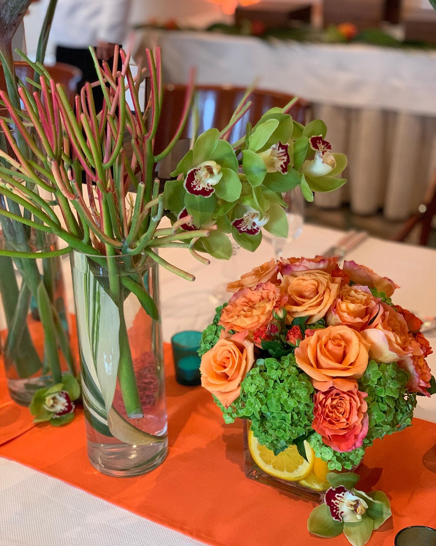 Tropical table floral arrangement with orange roses, green hydrangeas, and green orchids on orange runner.