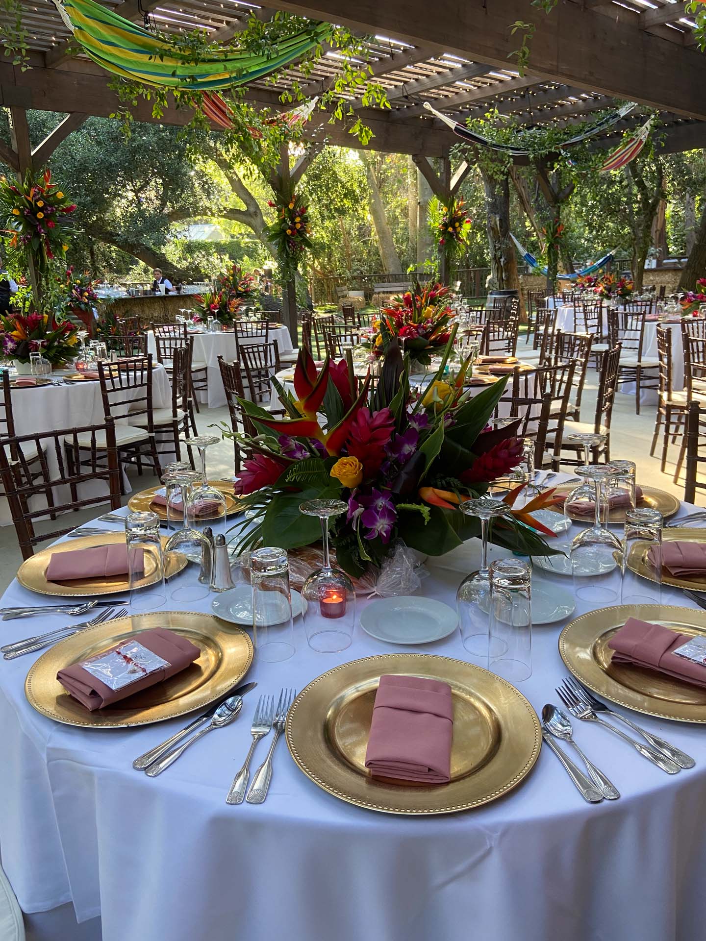 Outdoor dining area with round tables set with gold plates, pink napkins, floral centerpieces, and wooden chairs in a tropical theme.