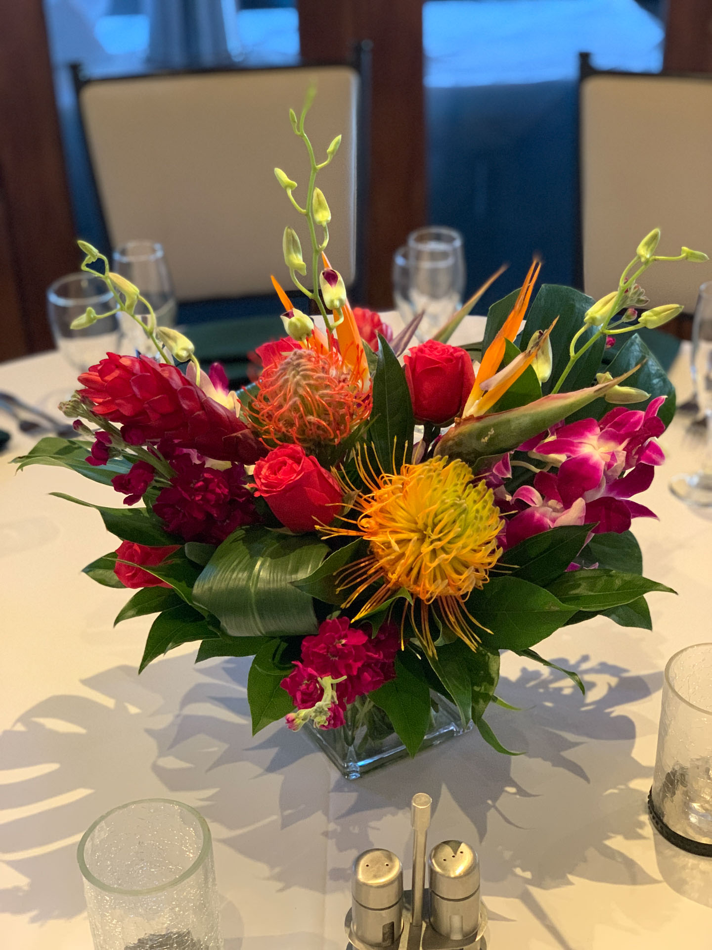 Colorful tropical floral centerpiece with red roses, yellow pincushion flowers, and tropical blooms on a white table.