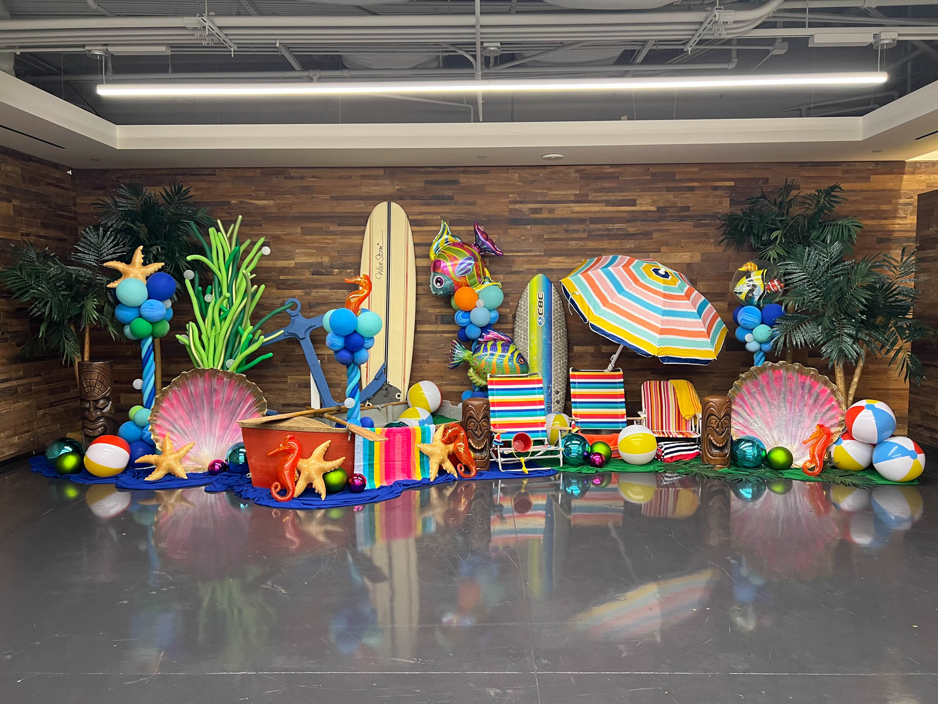 Indoor tropical beach-themed setup with surfboards, beach chairs, colorful umbrellas, starfish, balloons, and palm plants.
