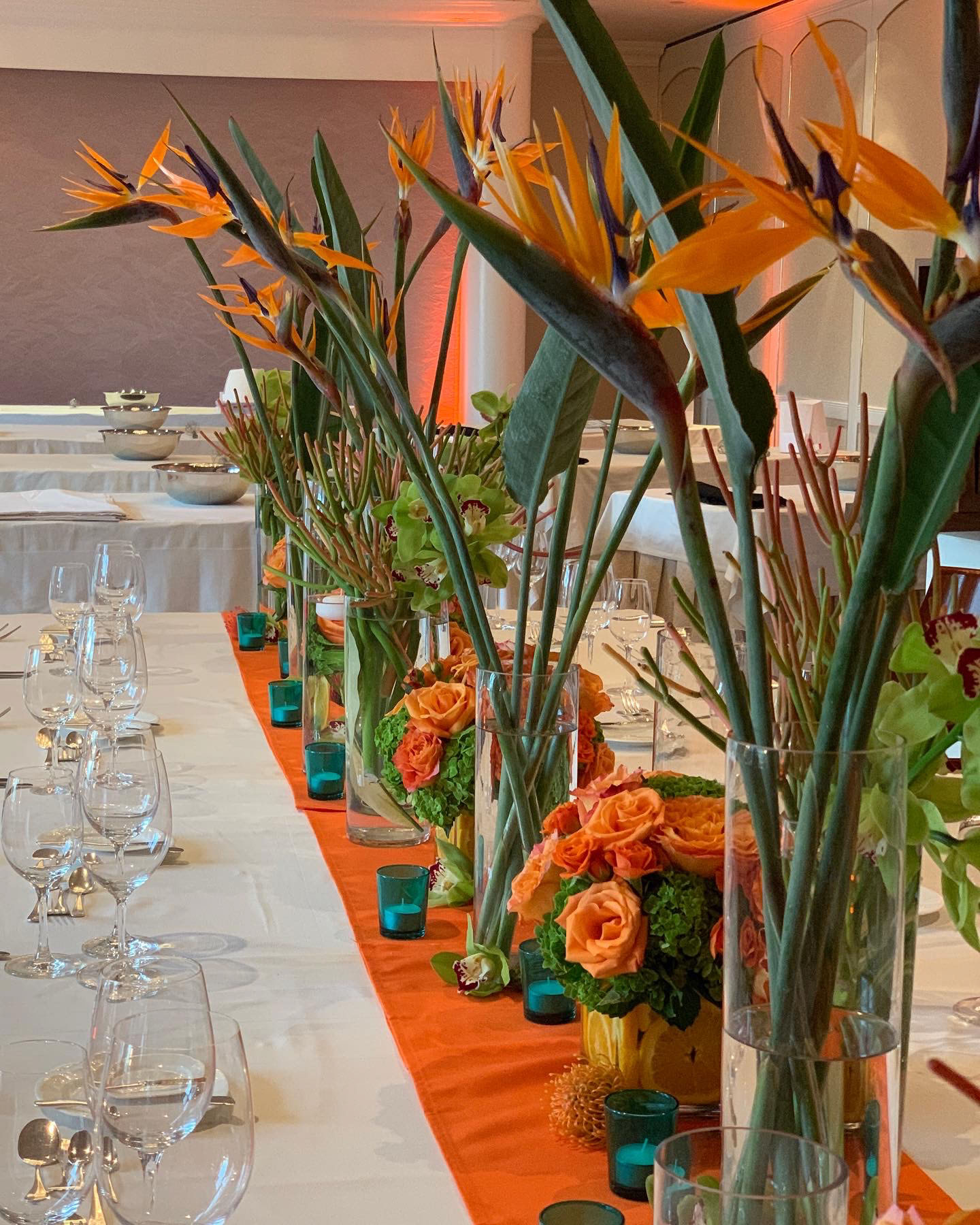 Long dining table with orange runner, tall bird of paradise flowers, orange roses, green orchids, and glassware.