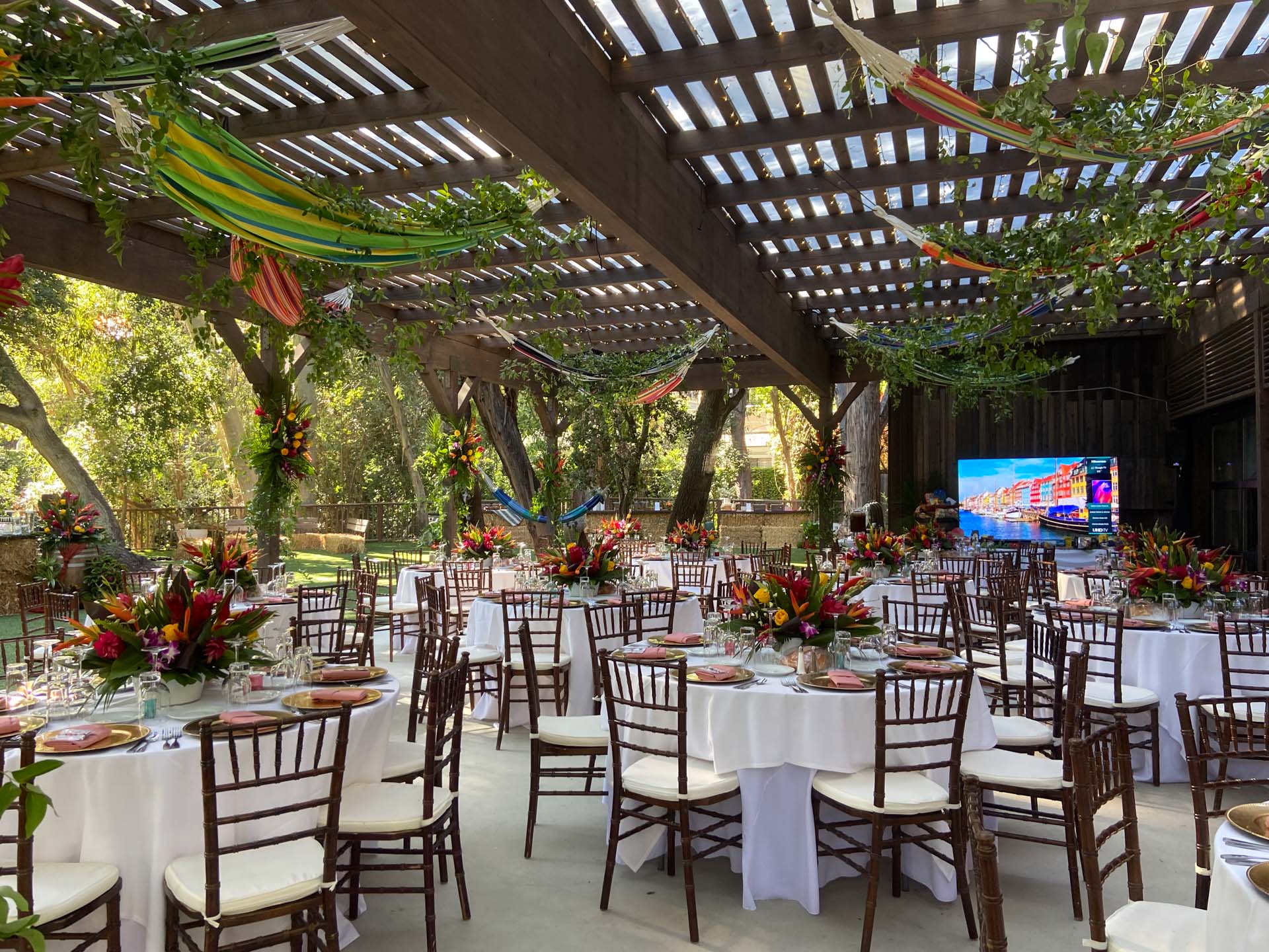 Outdoor event space with round tables, white tablecloths, colorful tropical floral centerpieces, and hammocks overhead in a tropical theme.