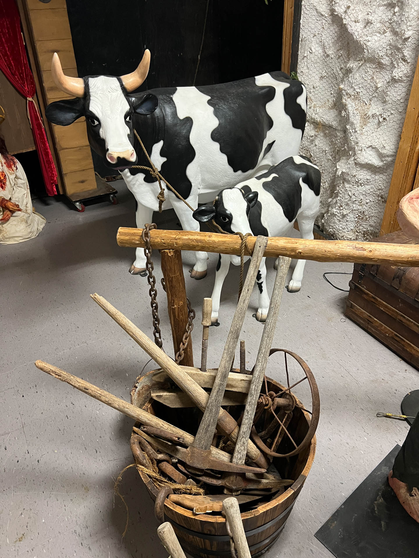 Two black and white cow statues behind a wooden bucket filled with antique farming tools indoors.