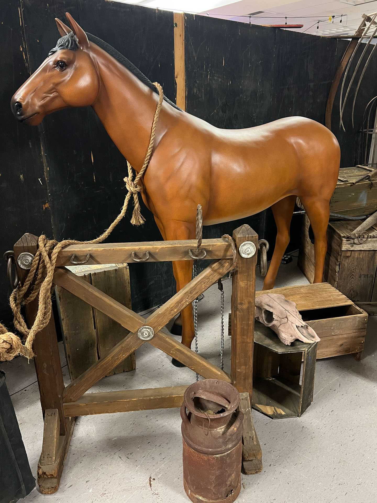 Life-size brown horse statue beside wooden crates, a skull, rustic farm tools, and a milk can.