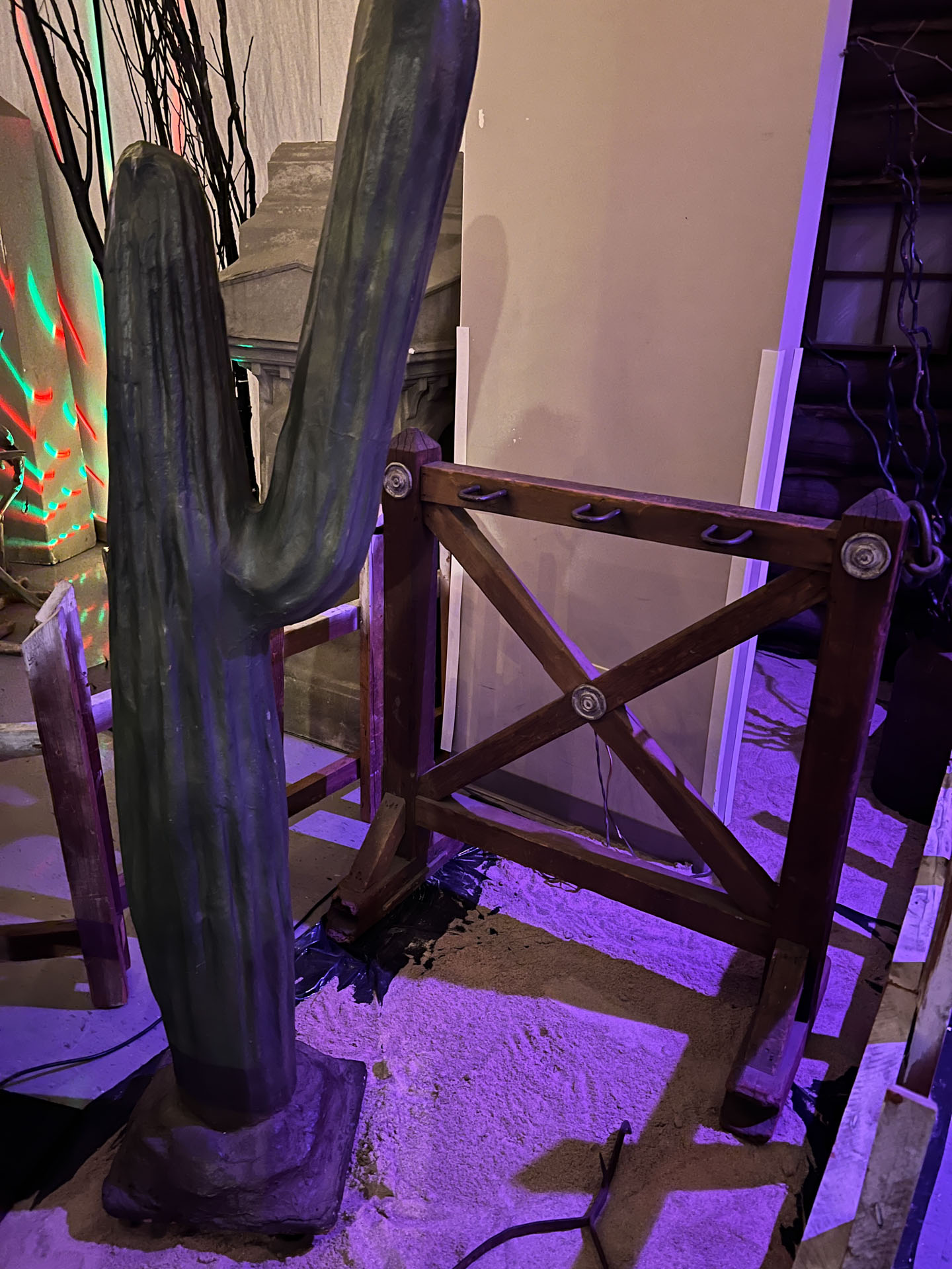 Artificial tall cactus next to wooden fence on sandy floor with purple lights.