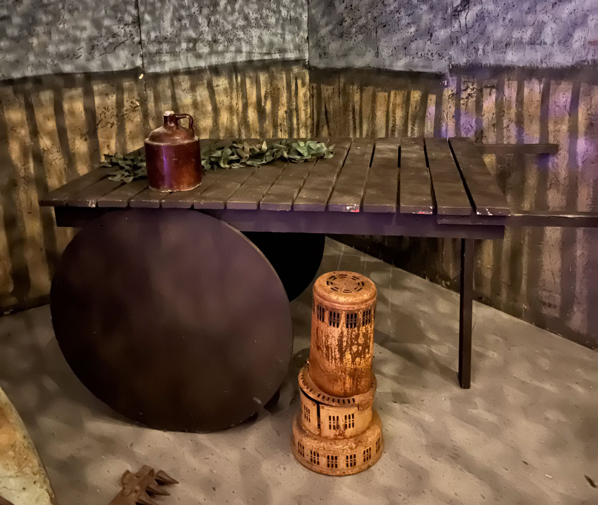 Rustic wooden table with a jug, greenery on top, and a rusty metal heater below in a dim room.