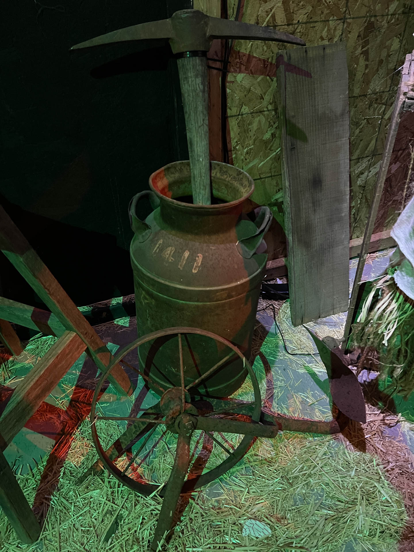 Rusty metal milk can with a wooden-handled pickaxe inside, surrounded by old tools and hay.