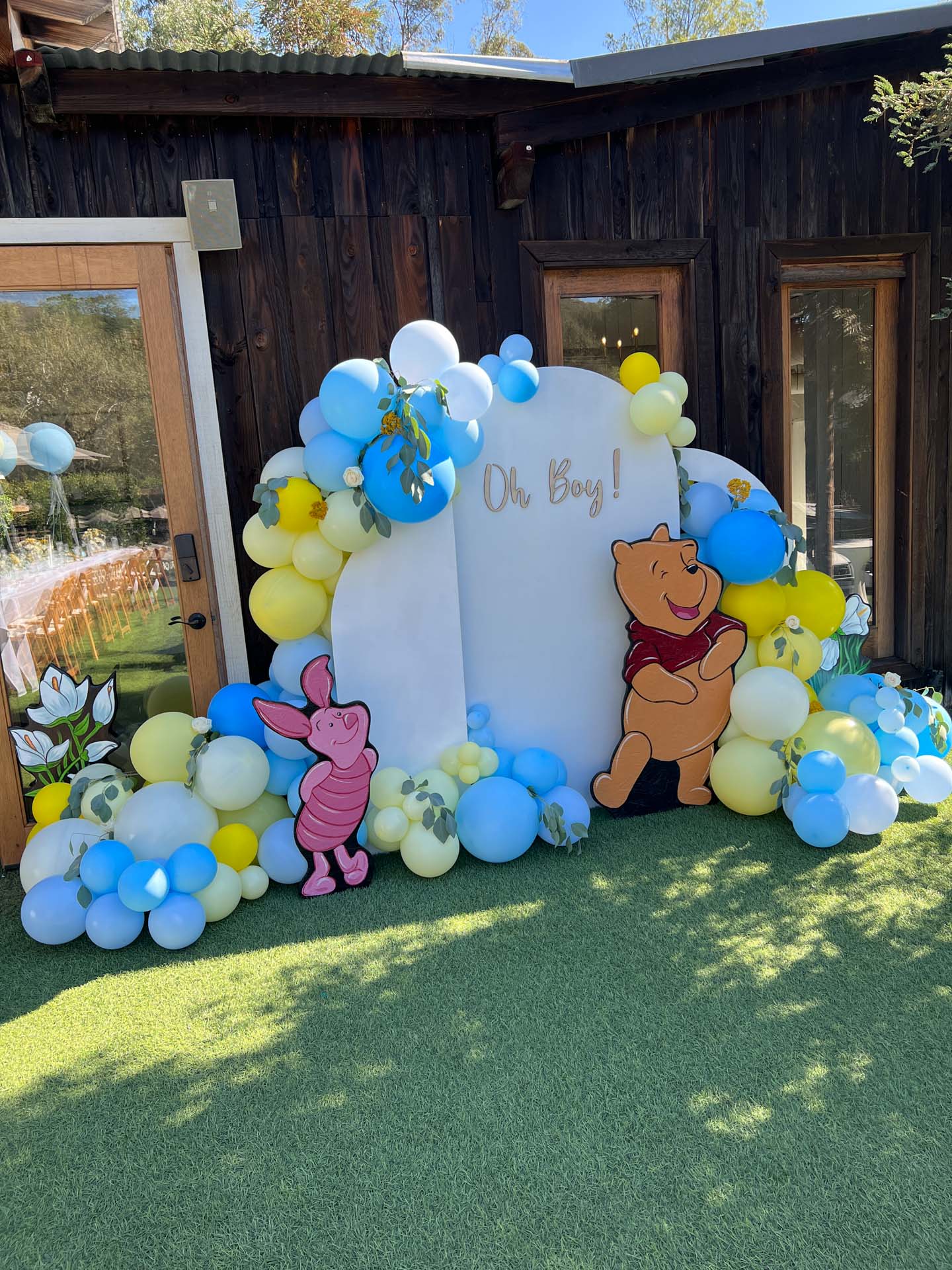 Winnie the Pooh and Piglet cutouts with blue, yellow, and white balloons surround a sign on a backdrop reading 'Oh Boy!'