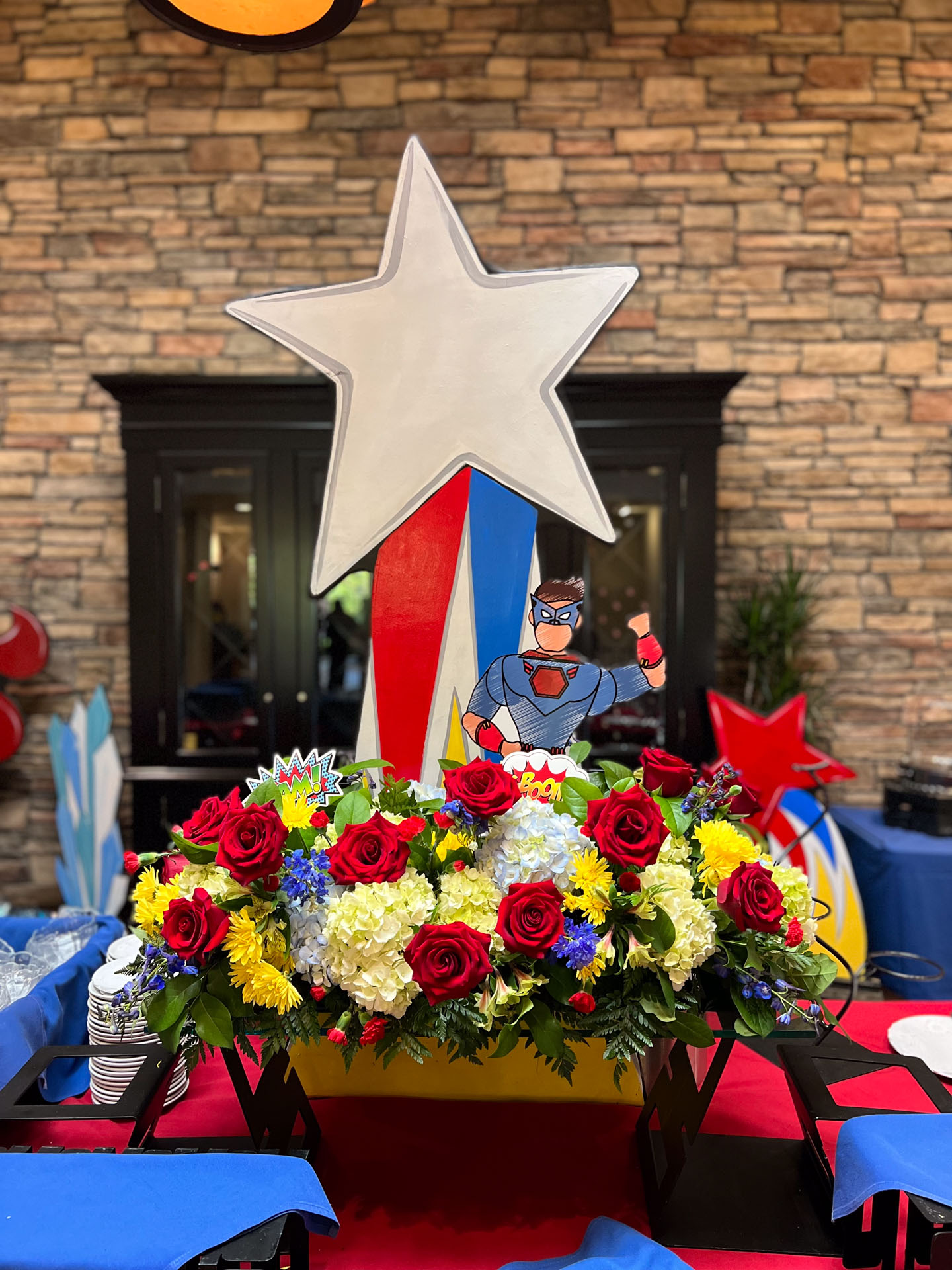 Table centerpiece with red roses, yellow and white flowers, a superhero cutout, and a large white star.