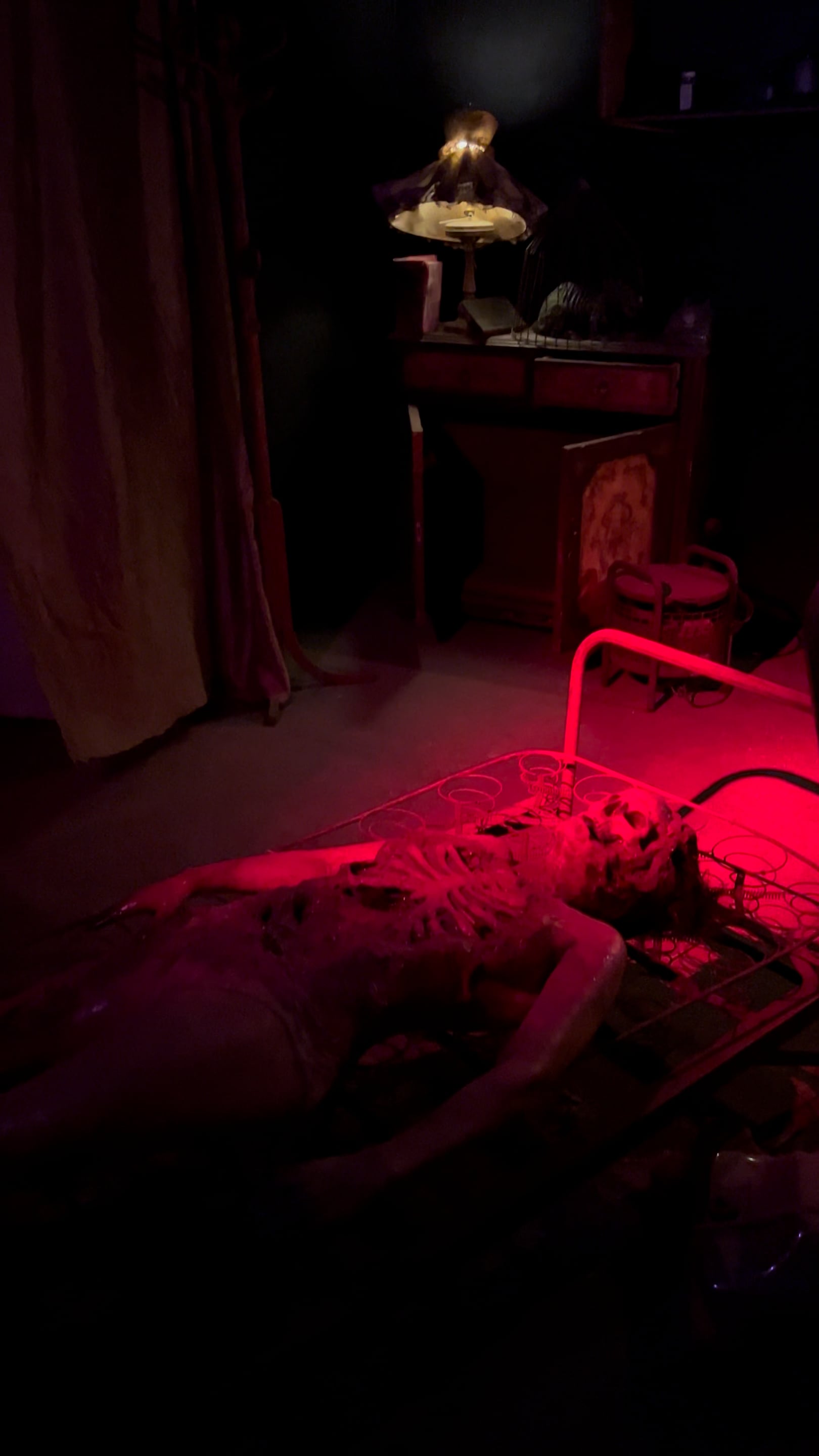 Dimly lit room with a skeleton-like figure lying on a bed frame under red light.