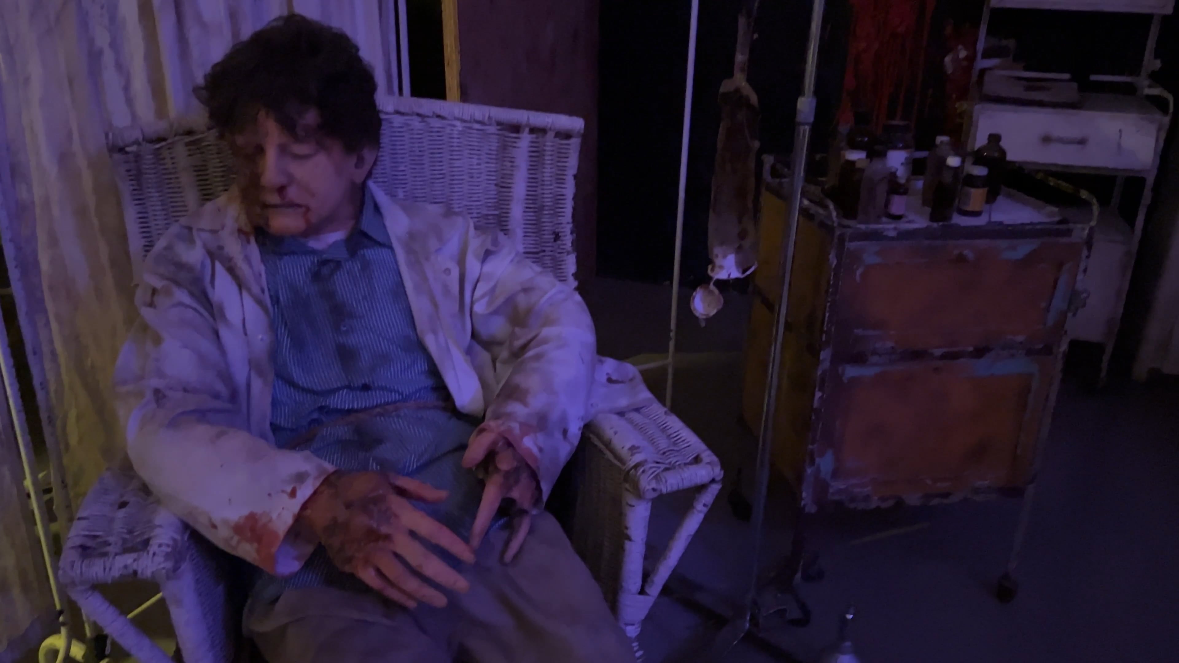 Man with boils in a blood-stained coat slumped in a wicker chair inside a dim room with medical supplies nearby.