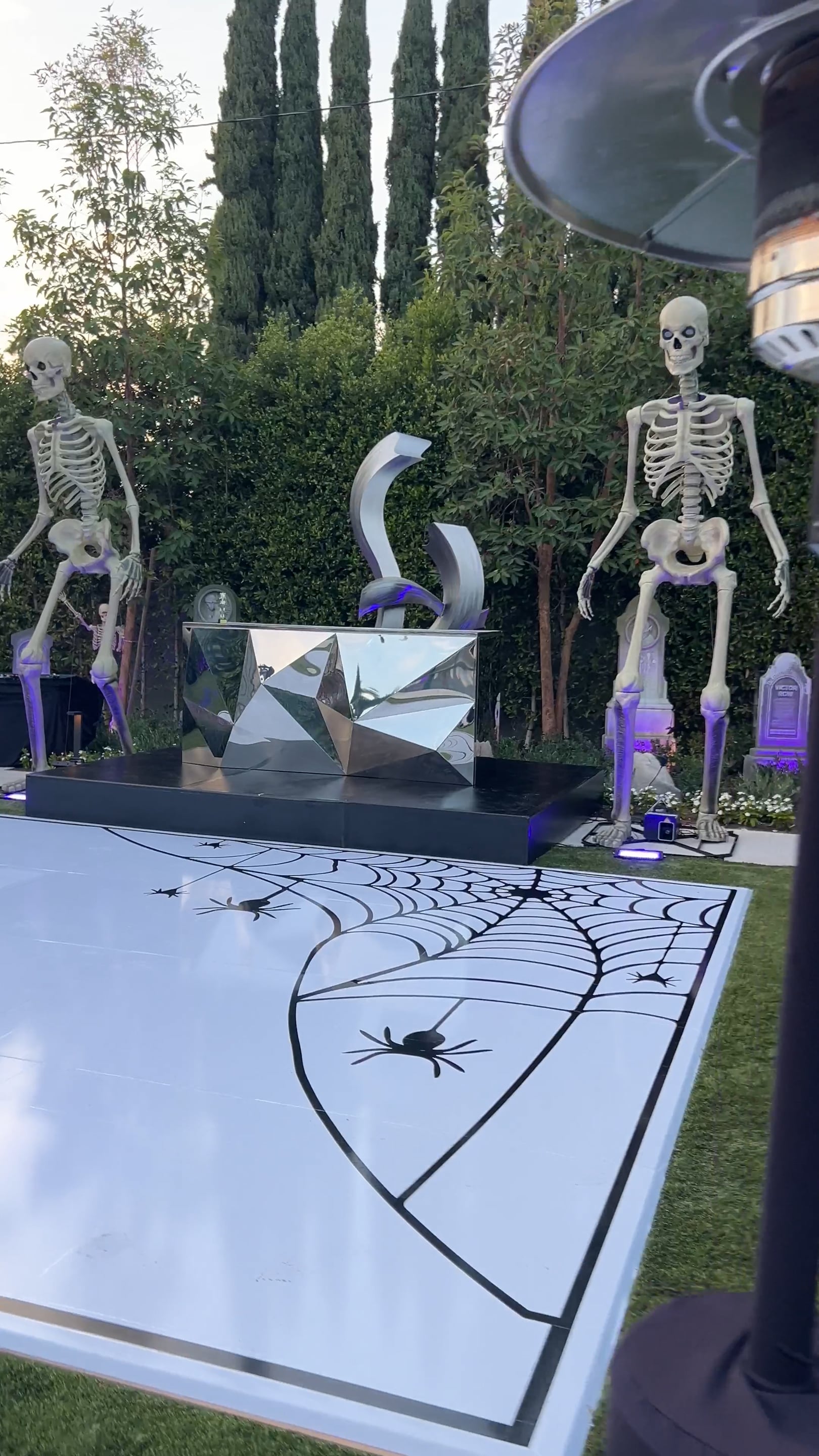 Outdoor Halloween party setup with two skeletons, tombstones, and a white dance floor with black spiderweb art.