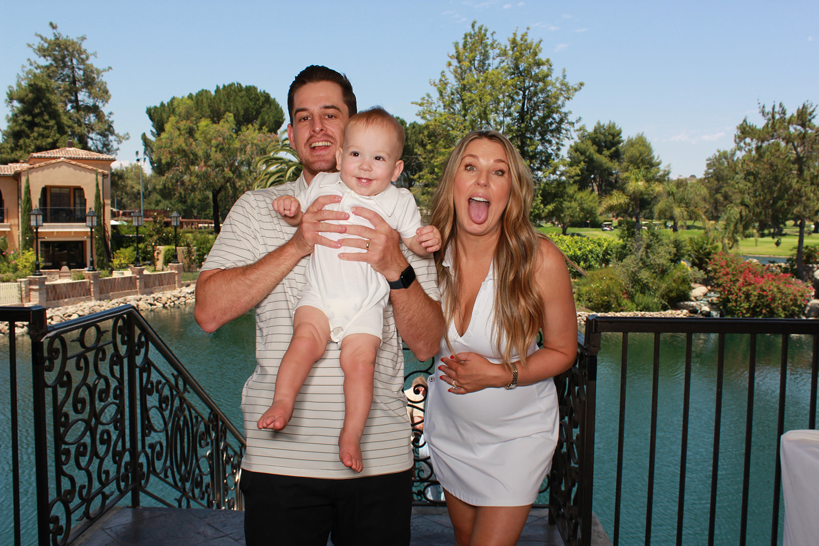 Man holding a smiling baby beside a woman making a playful face outdoors near a pond and landscaped grounds.