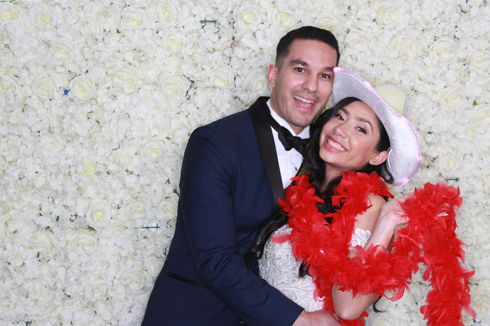 Couple posing in formal attire in front of a white flower wall, with the woman wearing a silver cowboy hat and red feather boa.