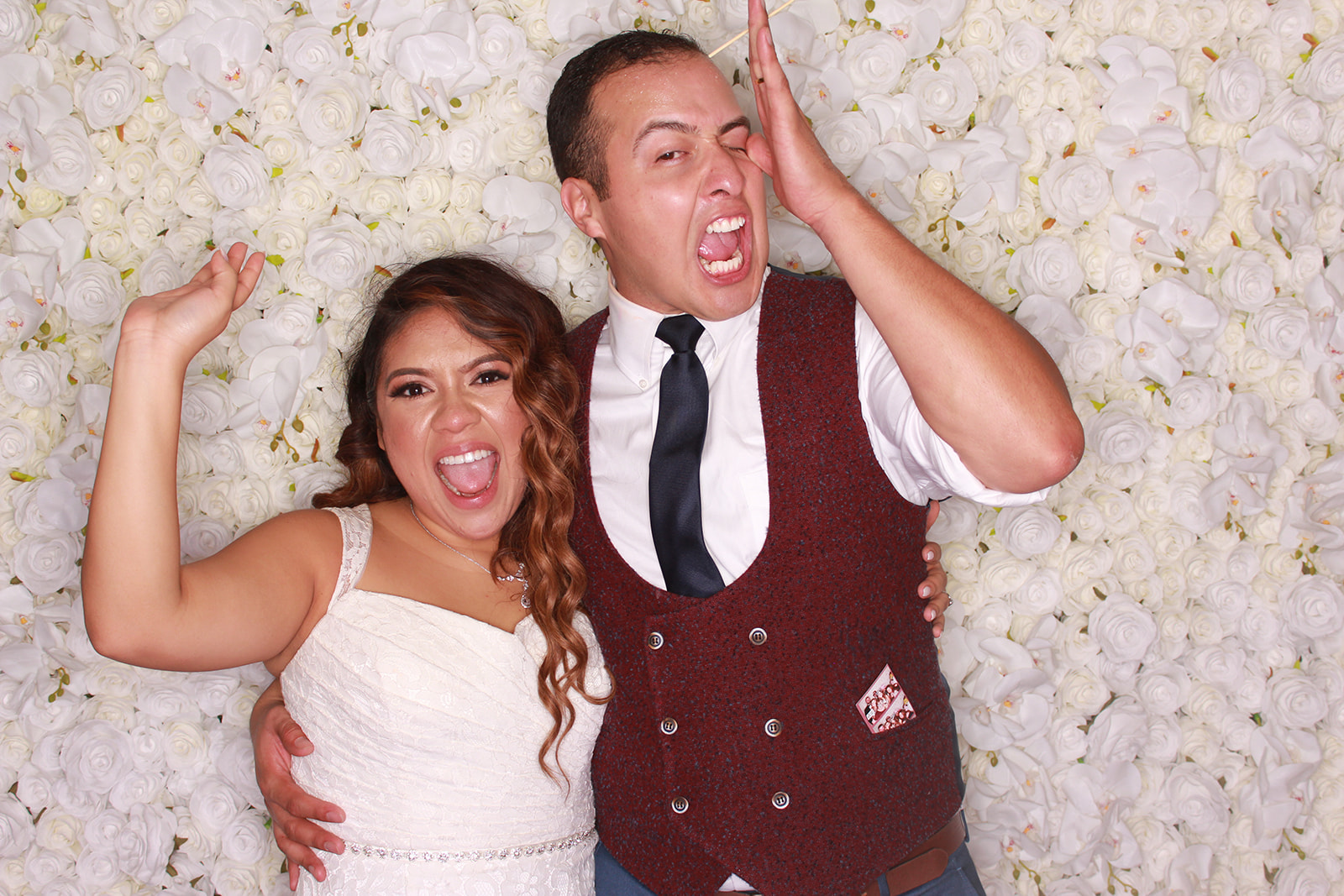Couple playfully posing in front of a white floral wall, with the woman raising one hand toward the man's face.