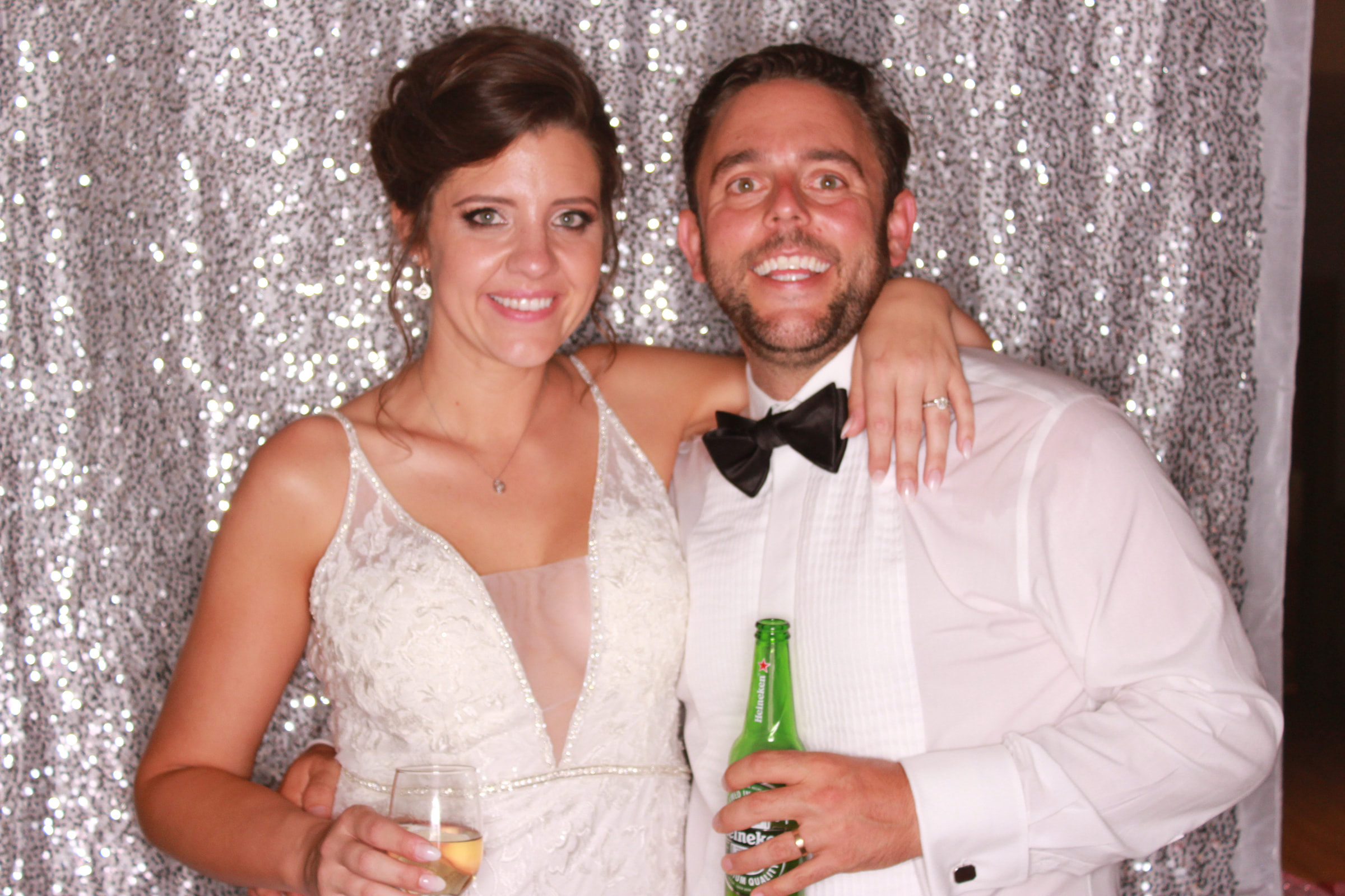 Bride and groom smiling together in front of a silver sequin backdrop, with the groom holding a beer bottle