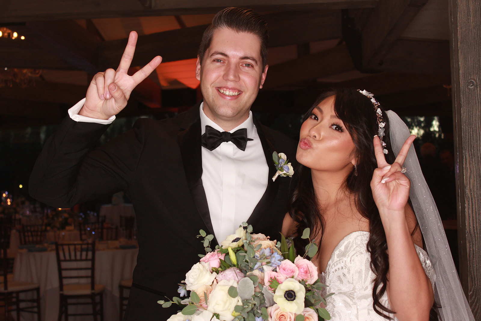 Bride and groom posing with peace signs while holding a colorful bouquet at their wedding reception.