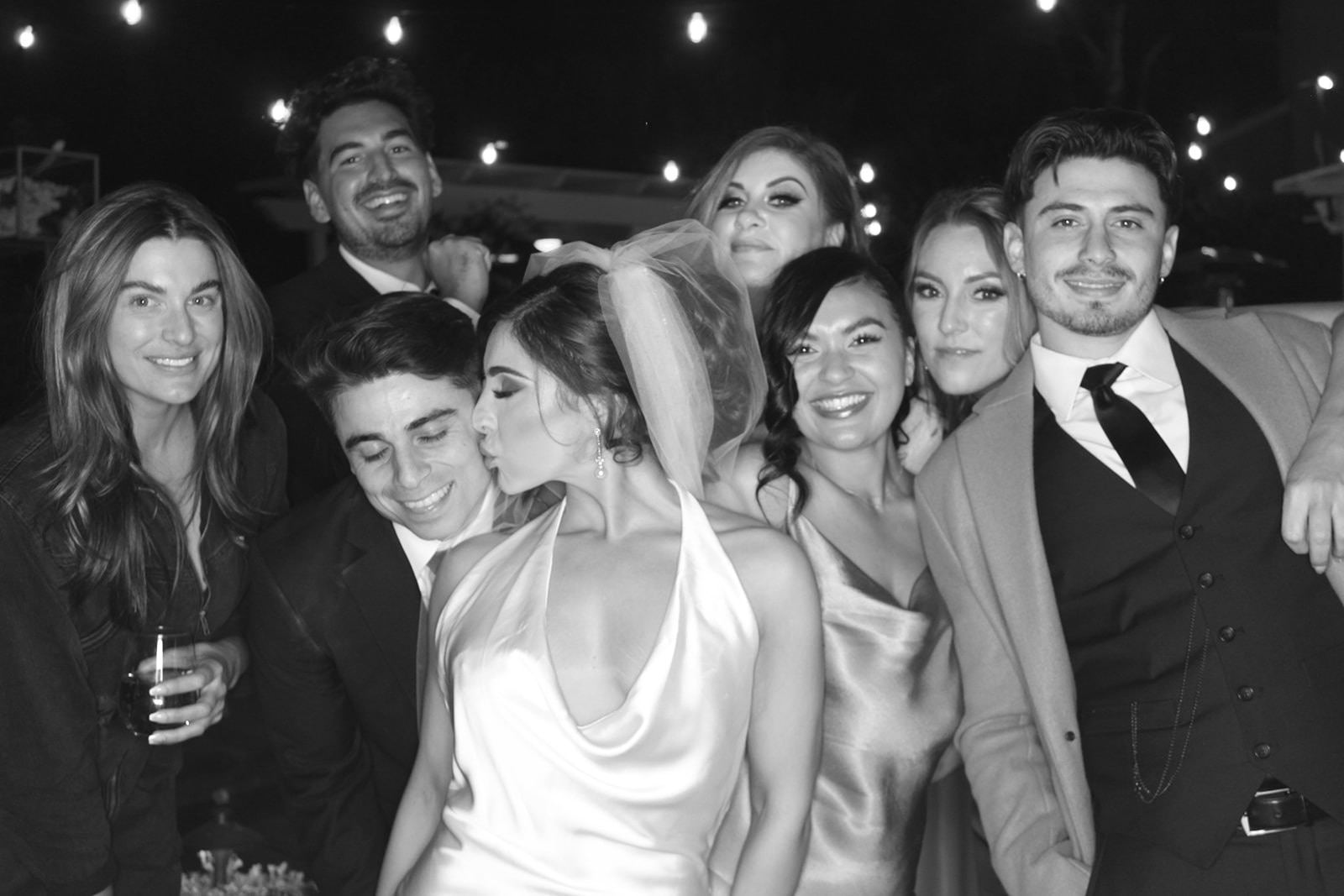 Black-and-white group portrait of wedding guests posing together outdoors, with the bride kissing the groom on the cheek.