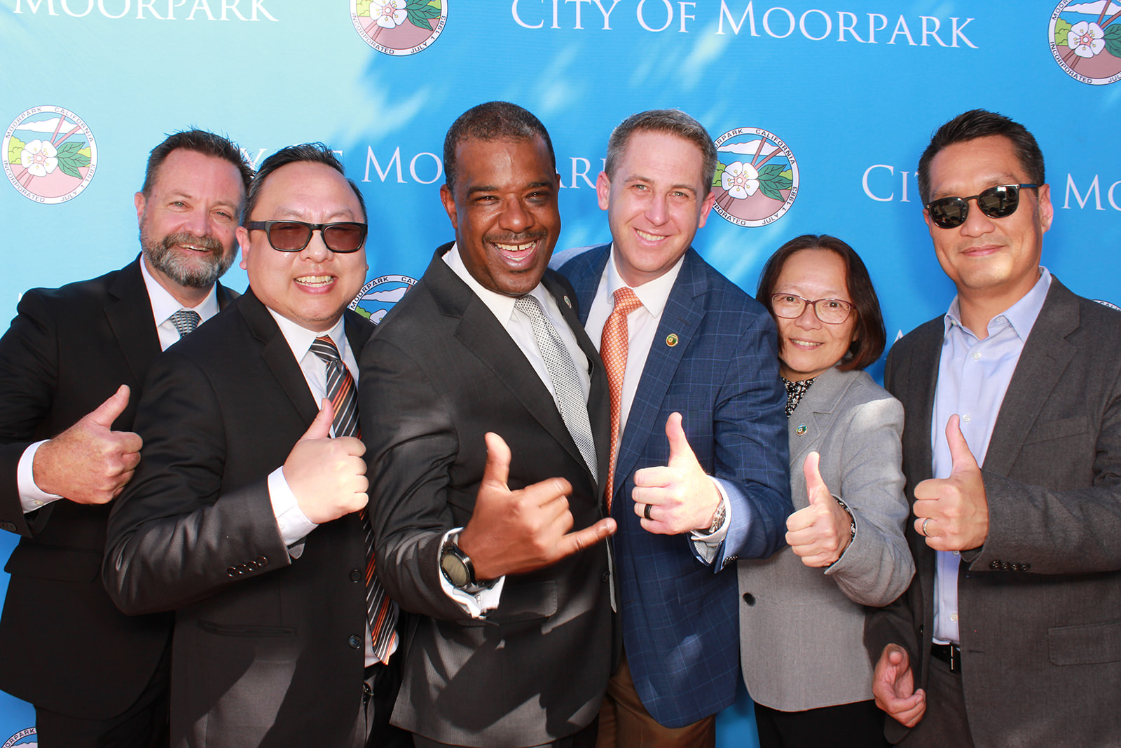 Group of six formally dressed adults posing with thumbs-up gestures in front of a blue City of Moorpark backdrop.