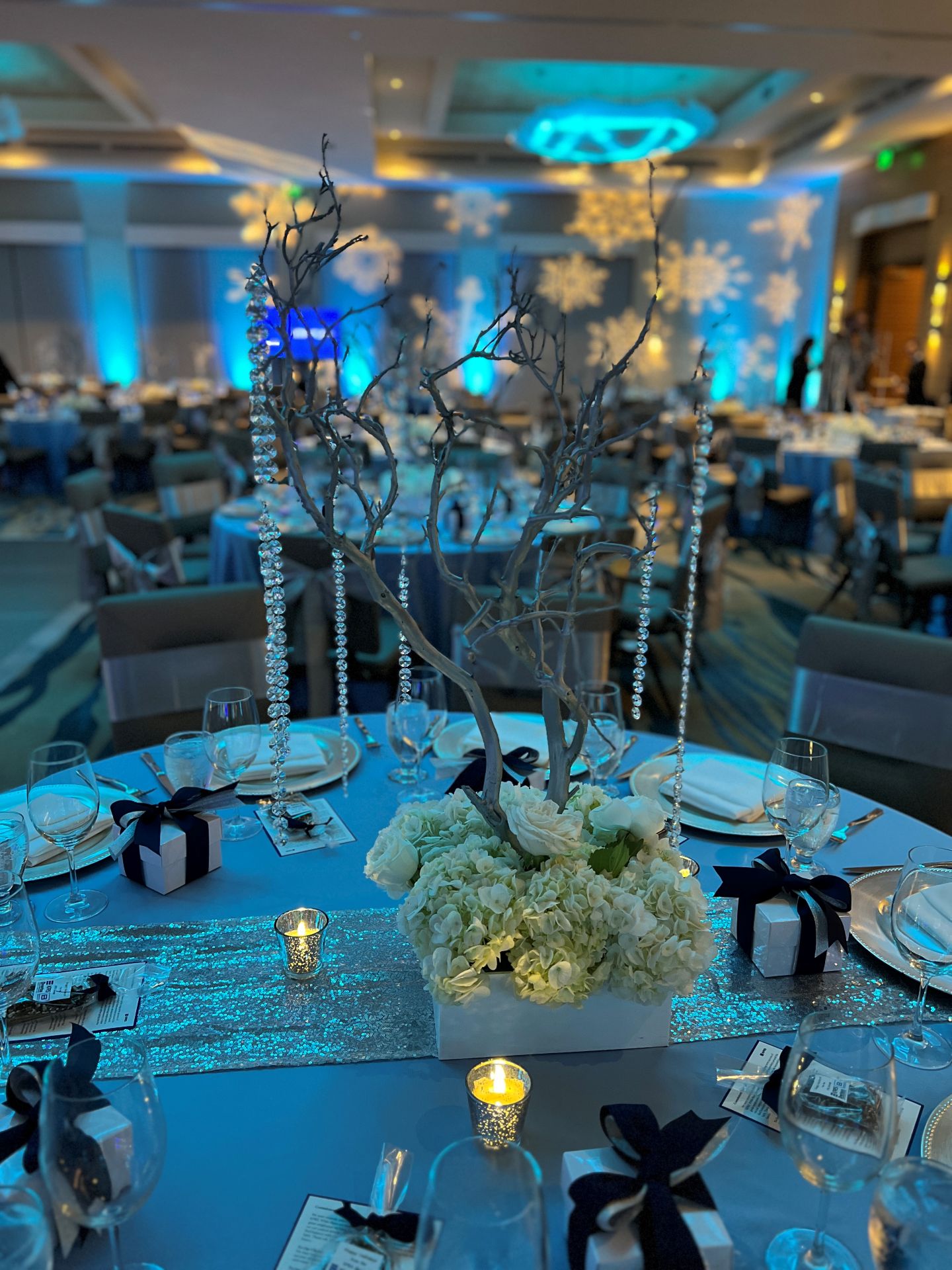 Elegant indoor reception table with white floral centerpiece, hanging crystal accents, candles, and blue ambient lighting.