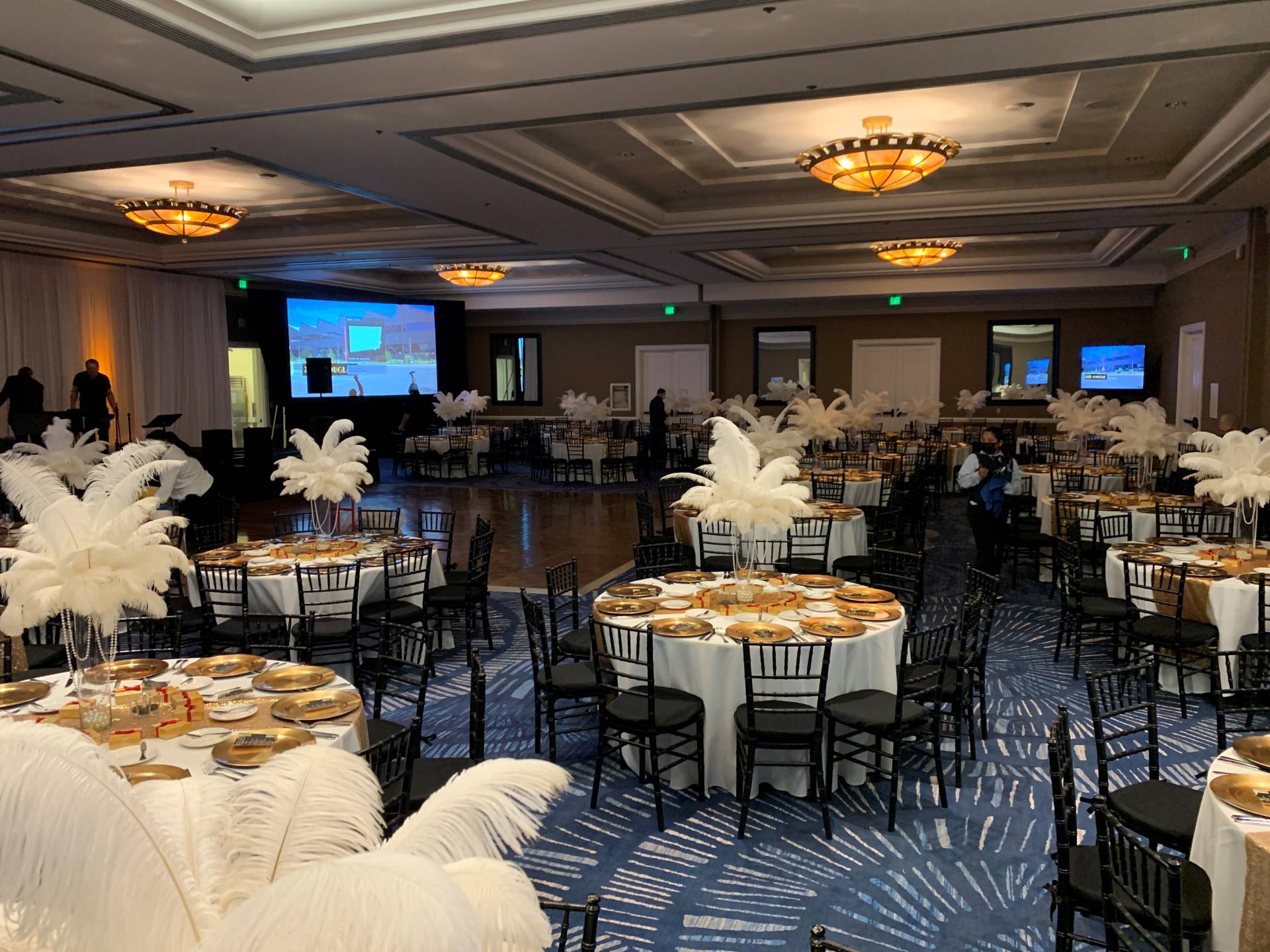 Large ballroom reception setup with round tables, gold place settings, black chairs, and tall white feather centerpieces.