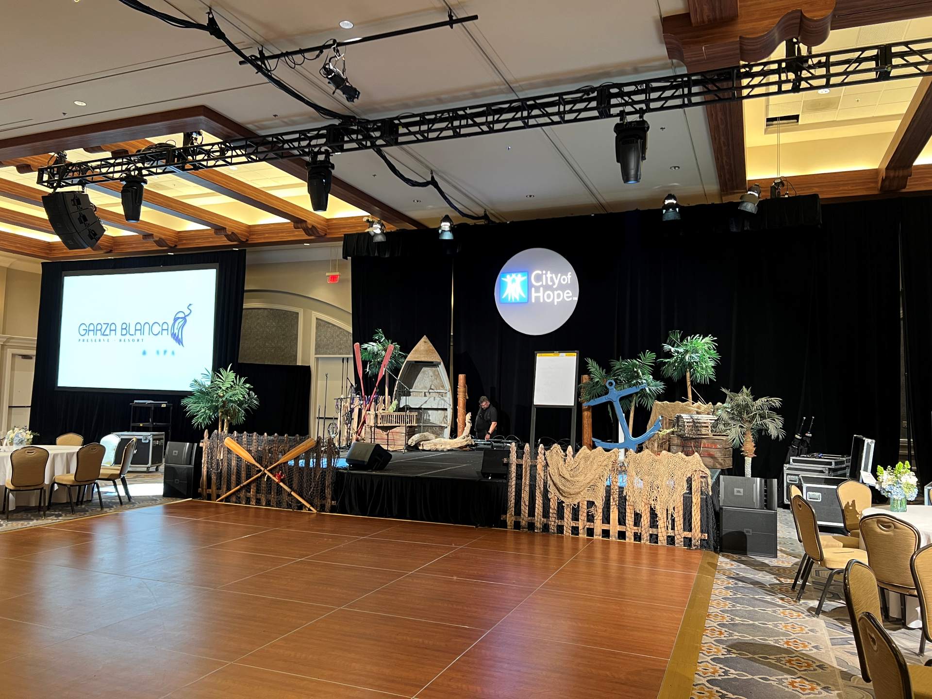 Event stage with tropical and nautical décor, lighting truss, projection screens, and a City of Hope backdrop.