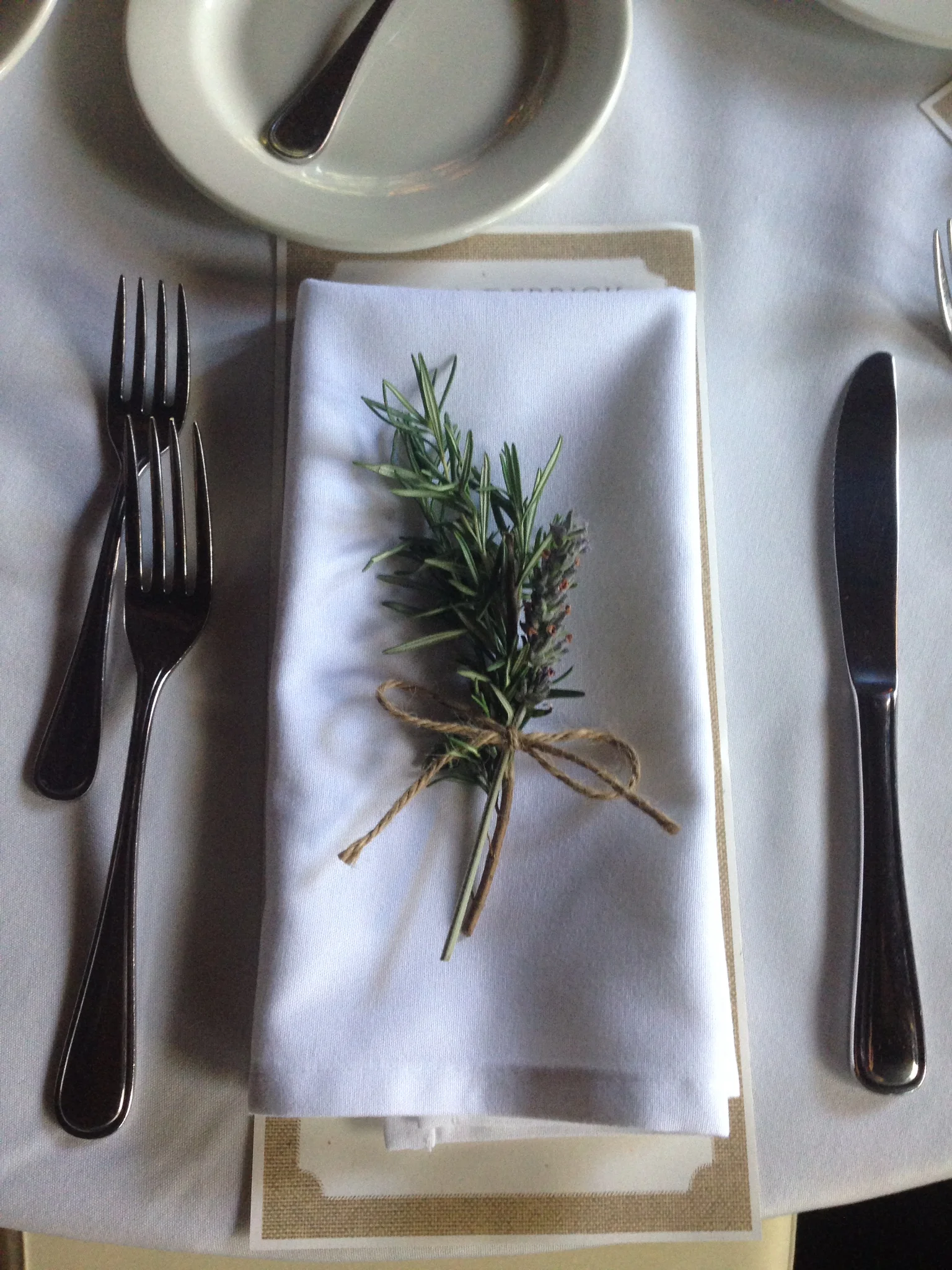 Formal place setting with a white napkin topped with tied sprigs of rosemary and lavender.
