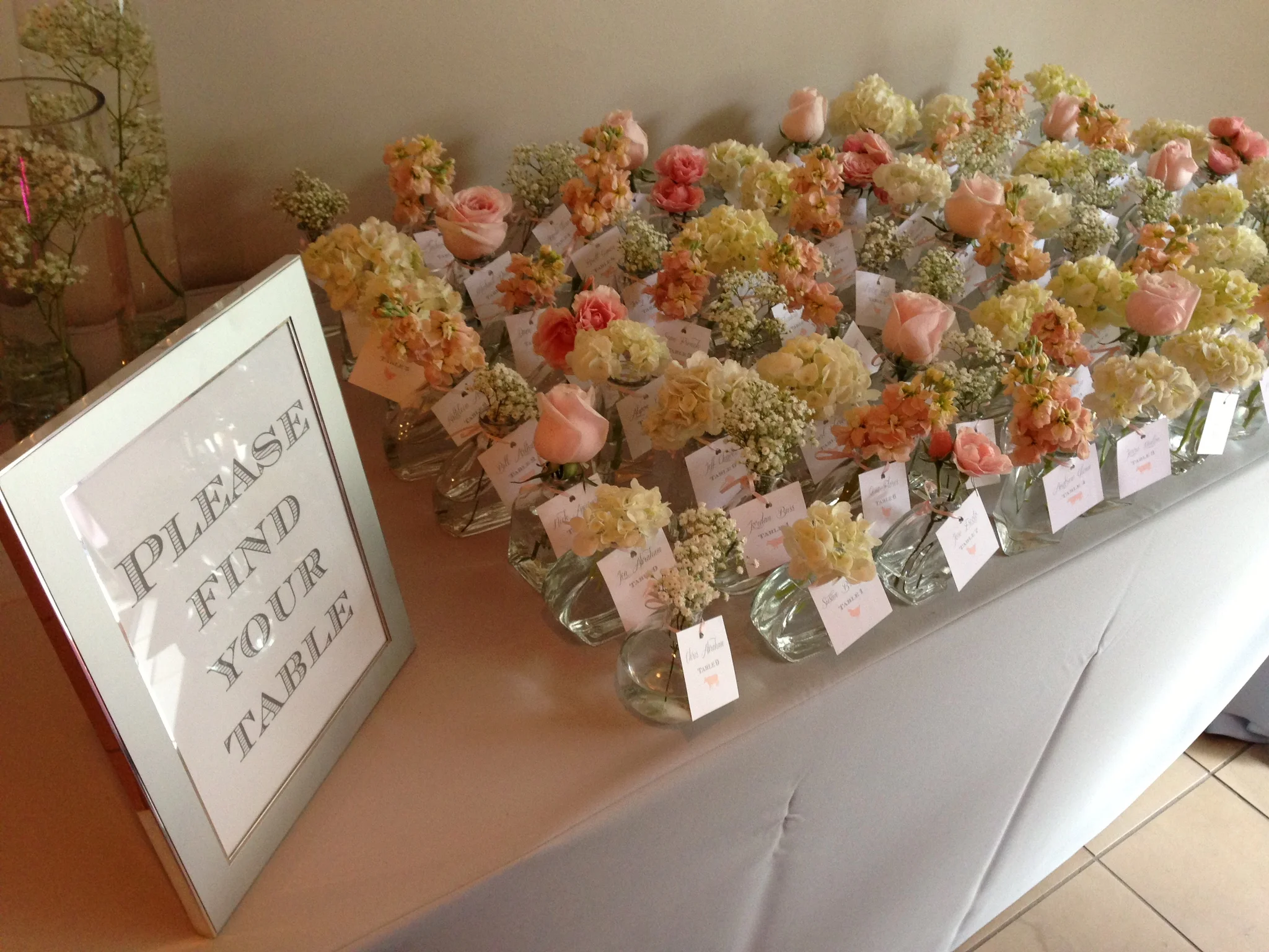 Wedding escort card display featuring small floral arrangements of pink roses, hydrangeas, and baby's breath in glass vases.