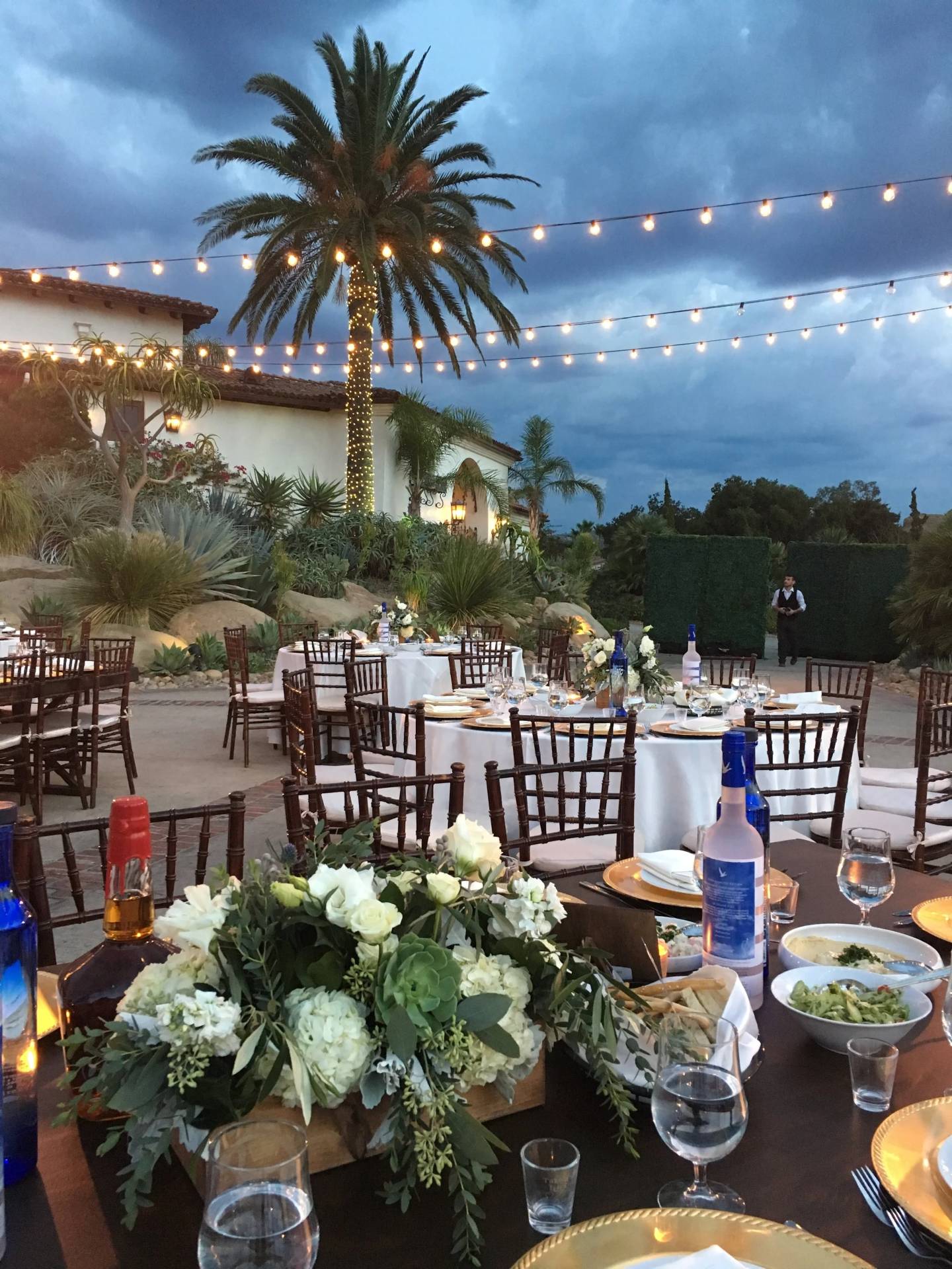 Outdoor reception tables with white floral centerpieces, gold place settings, wooden chairs, and string lights beneath a tall palm tree.