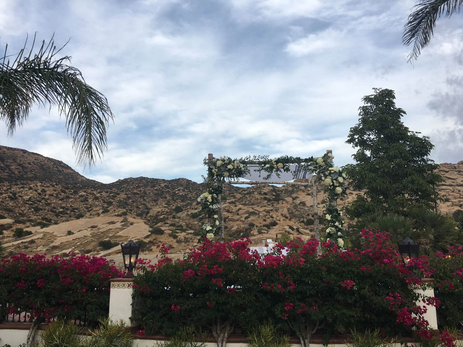 Outdoor wedding ceremony area with a flower-covered wooden arch overlooking rocky hills and bright bougainvillea.