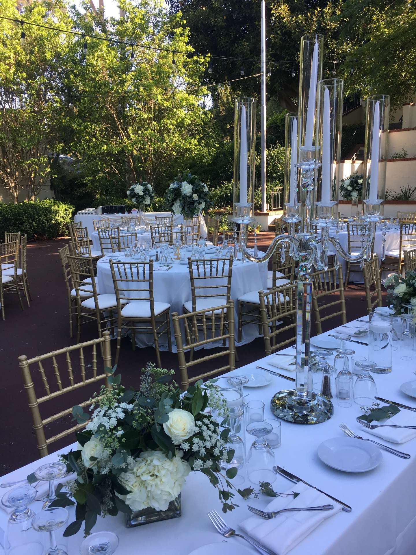Outdoor reception tables with white linens, gold chairs, white floral arrangements, and tall clear candleholders.