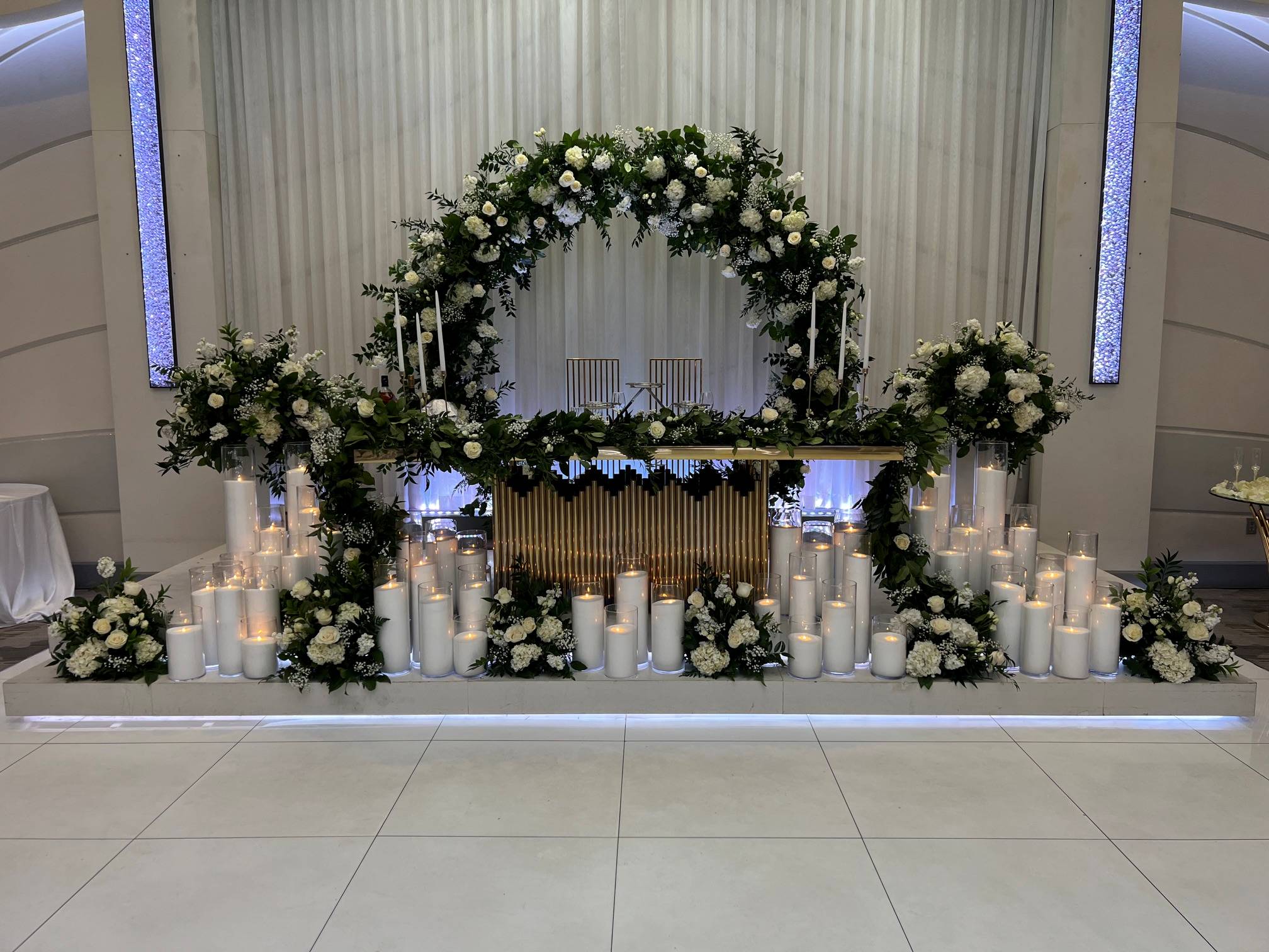 Elegant white and green floral arch surrounding a sweetheart table with lush greenery, white flowers, and glowing pillar candles.