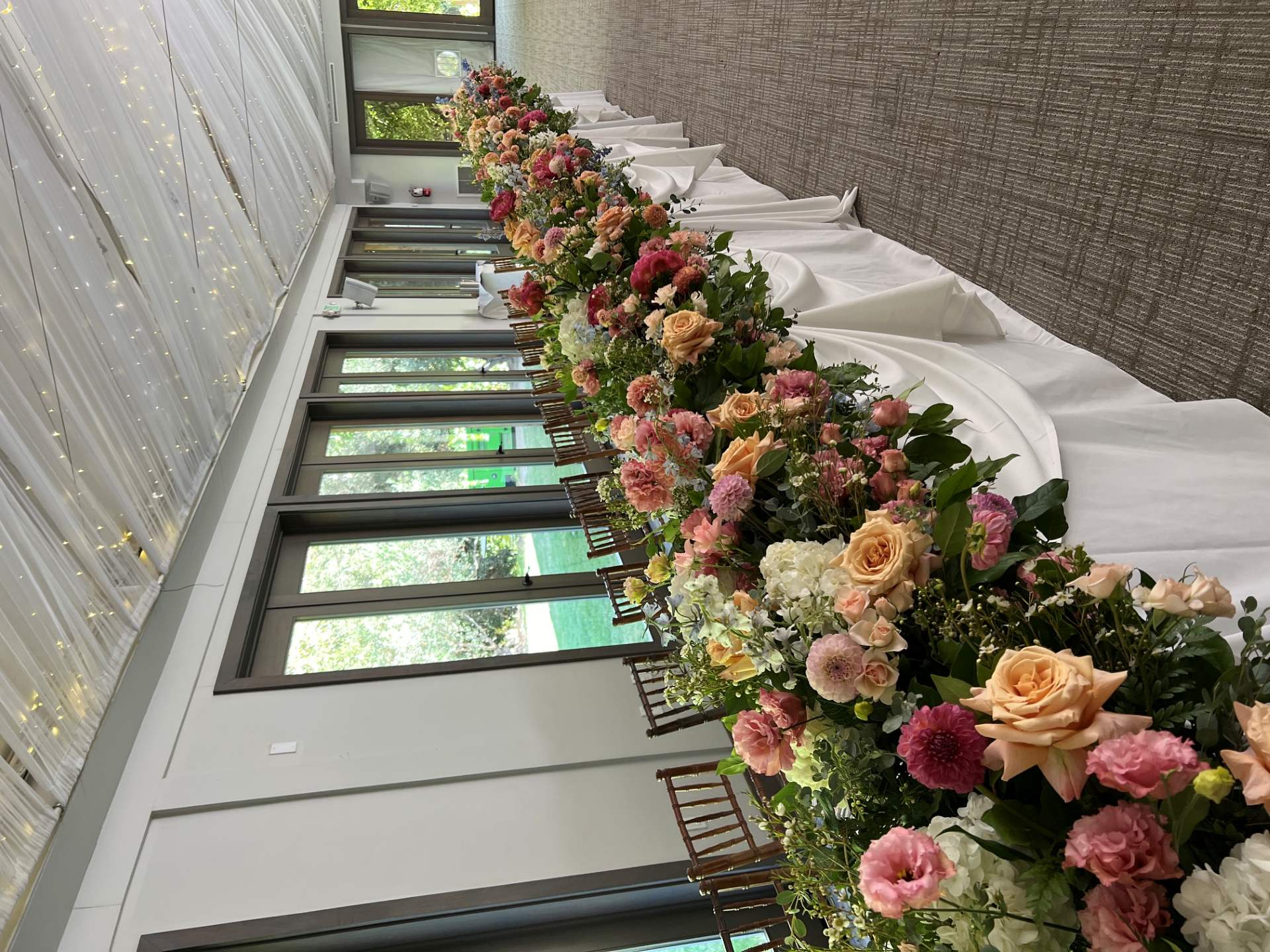 Long reception table lined with abundant pink, peach, white, and burgundy floral arrangements beneath a softly lit draped ceiling.