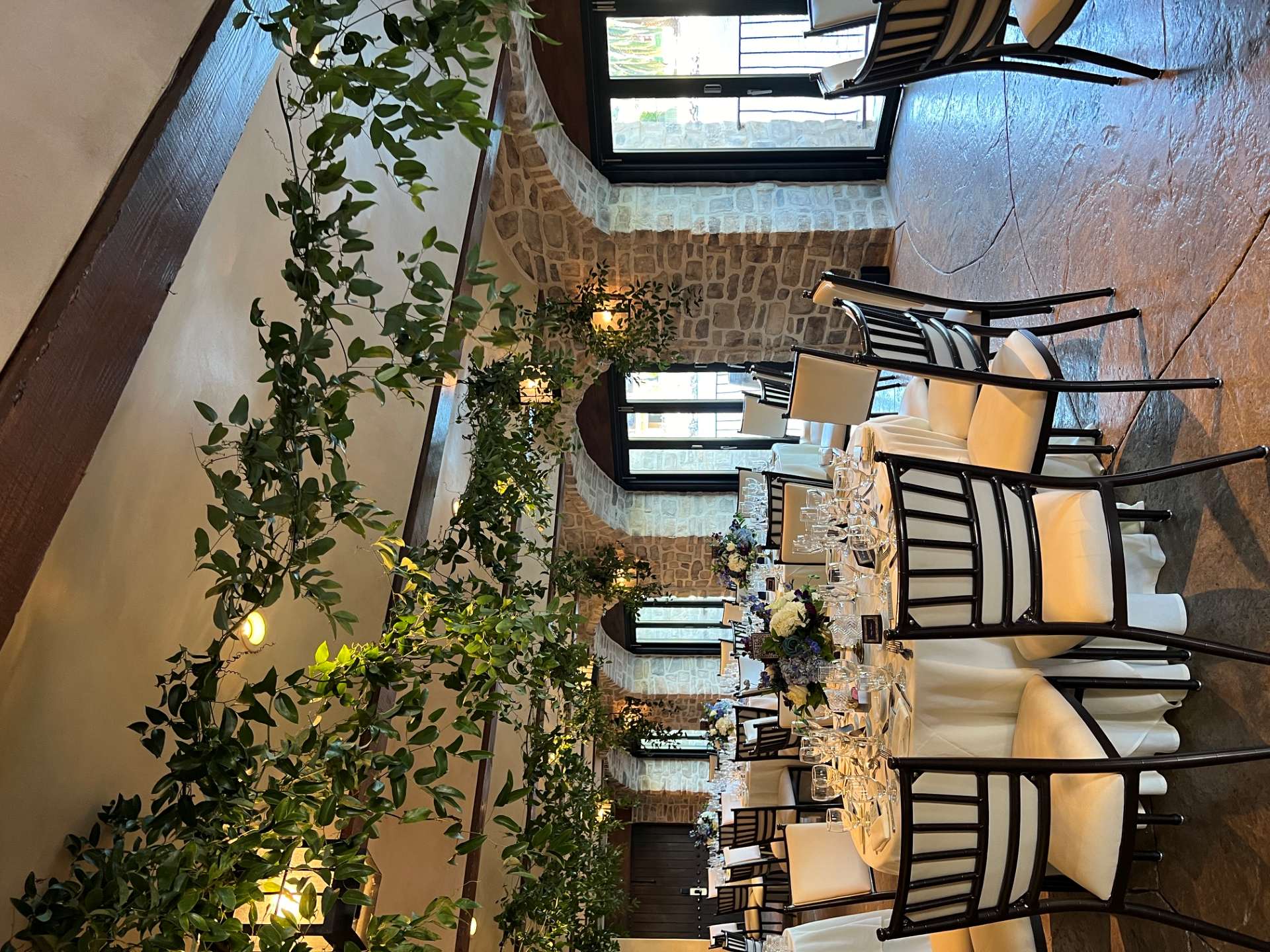 Indoor reception space with round dining tables, floral centerpieces, stone archways, and greenery draped across the ceiling.