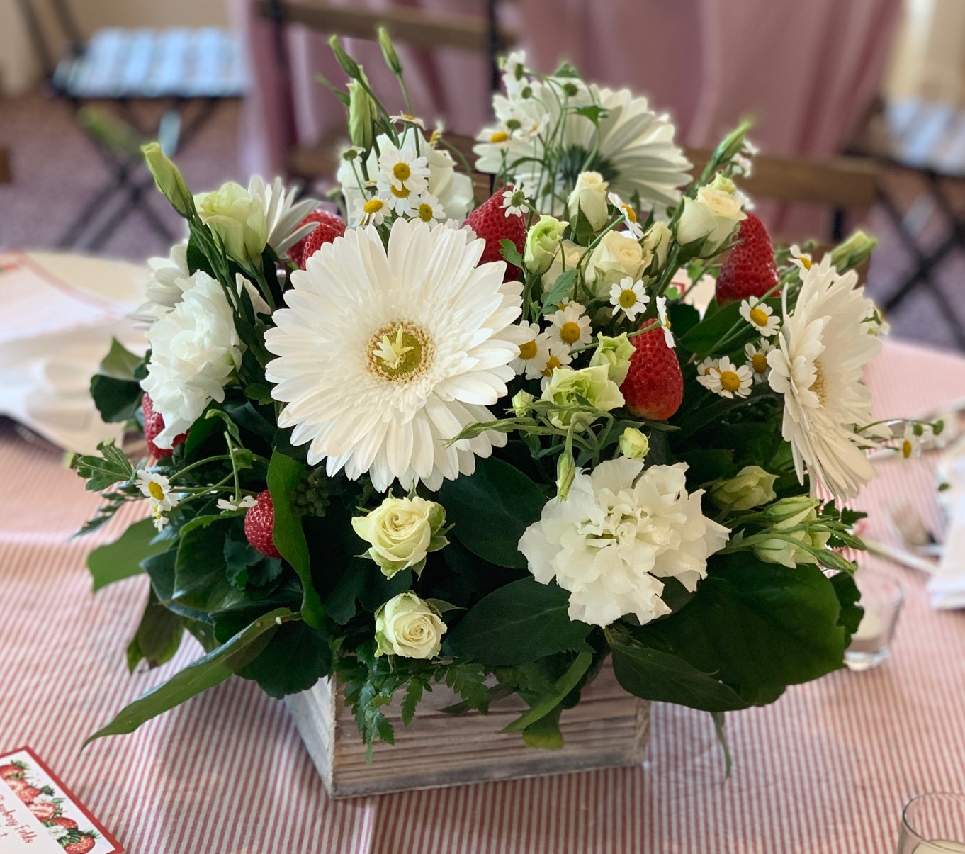 White floral centerpiece with gerbera daisies, roses, small daisies, greenery, and fresh strawberries.