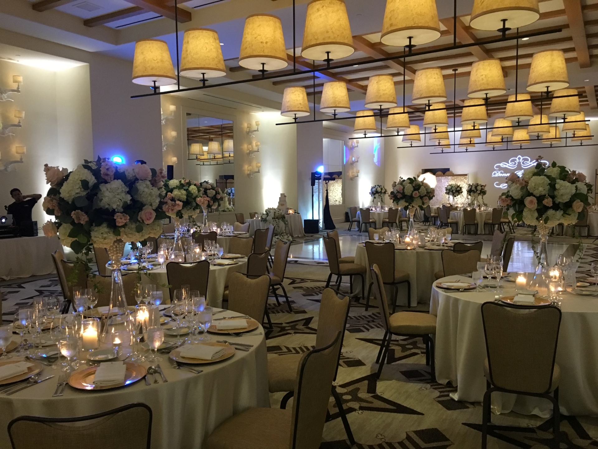 Elegant ballroom reception with tall white and blush floral centerpieces, candlelit tables, and warm hanging lights.