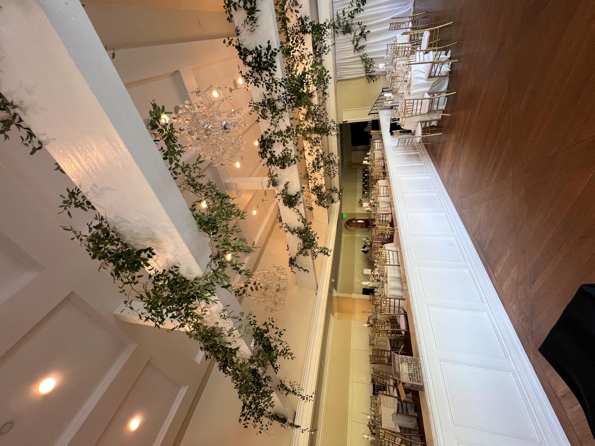 Wedding reception room with greenery cascading from ceiling beams, crystal chandeliers, round tables, and gold chairs.