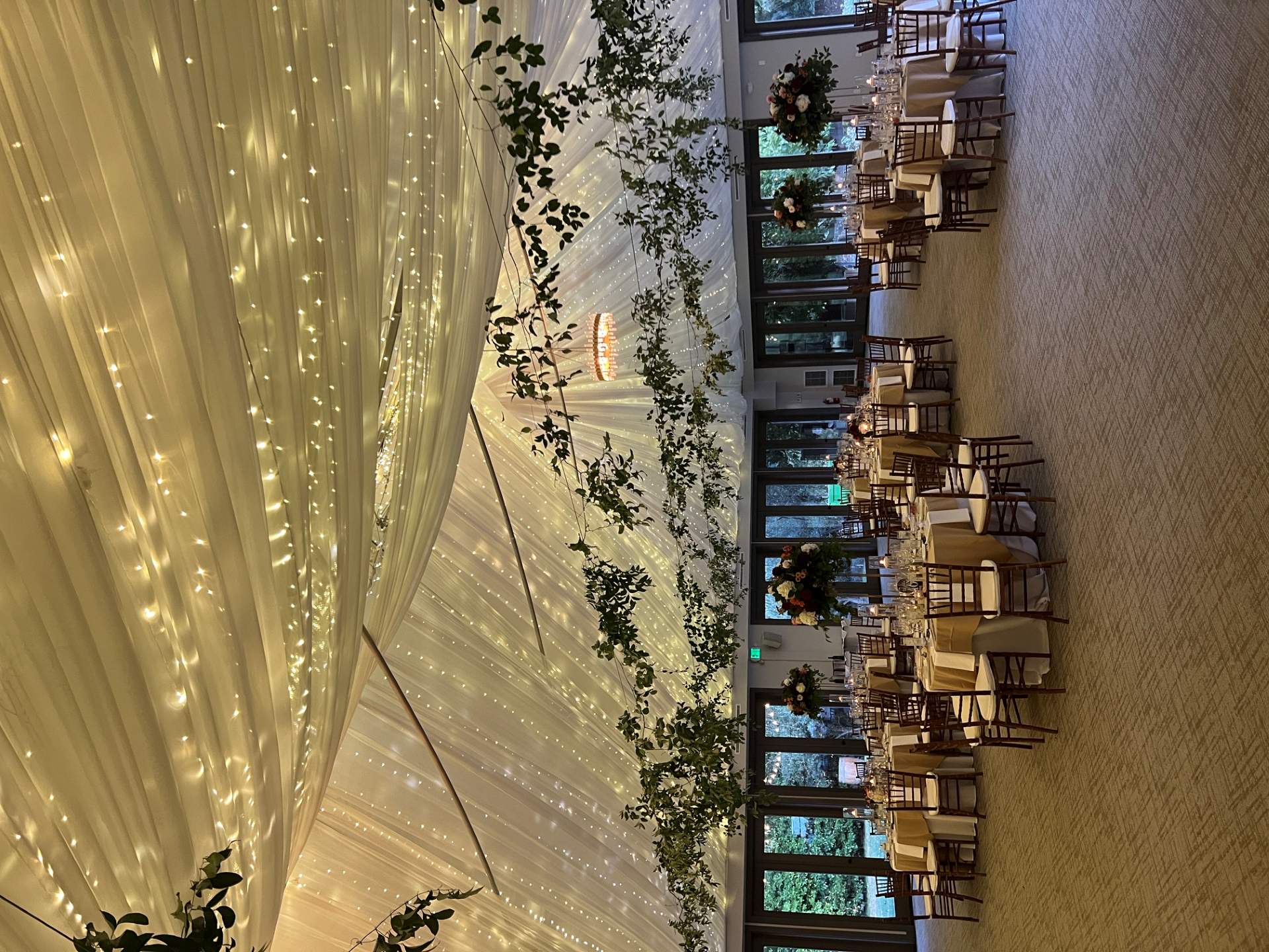 Reception space beneath a white draped ceiling filled with warm string lights and suspended greenery above decorated tables.