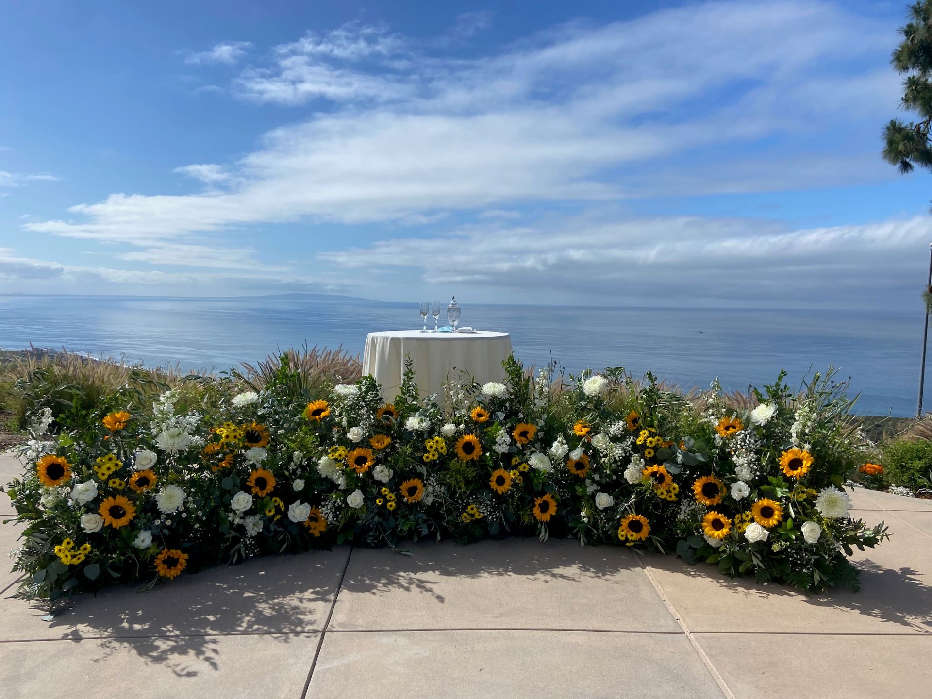 Large arrangement of sunflowers, white flowers, and greenery framing a small table overlooking the ocean.